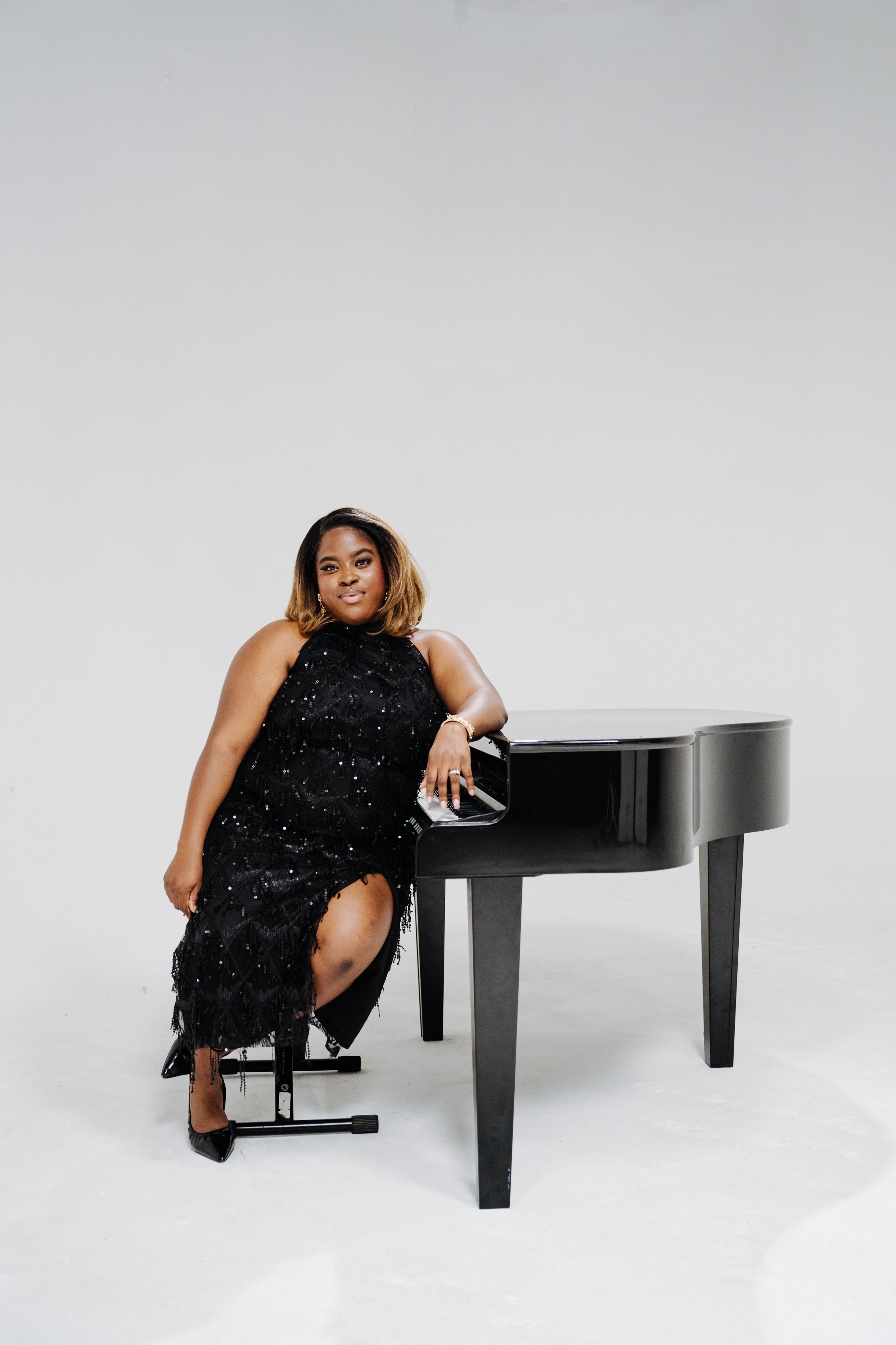 A woman dressed in a black, sequined dress is leaning on a black grand piano in a minimal white studio setting.