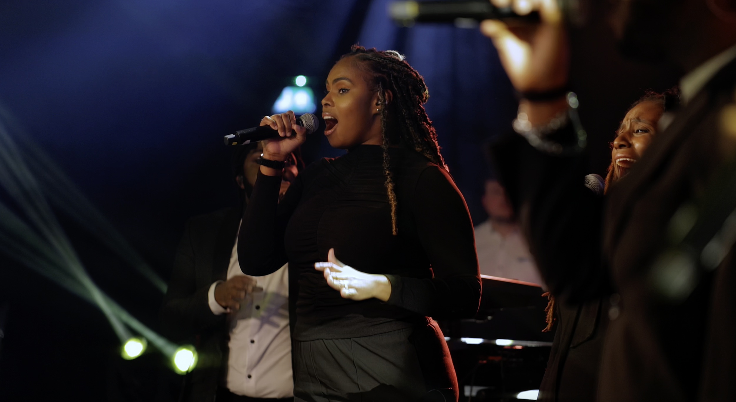 A woman singing into a microphone on stage with other singers in the background, illuminated by stage lighting.