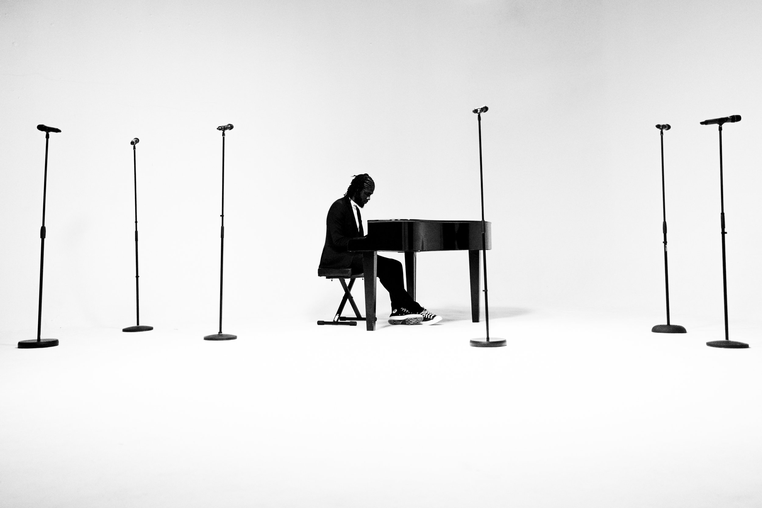 A person in a suit playing a grand piano on a white stage, surrounded by eight microphone stands.