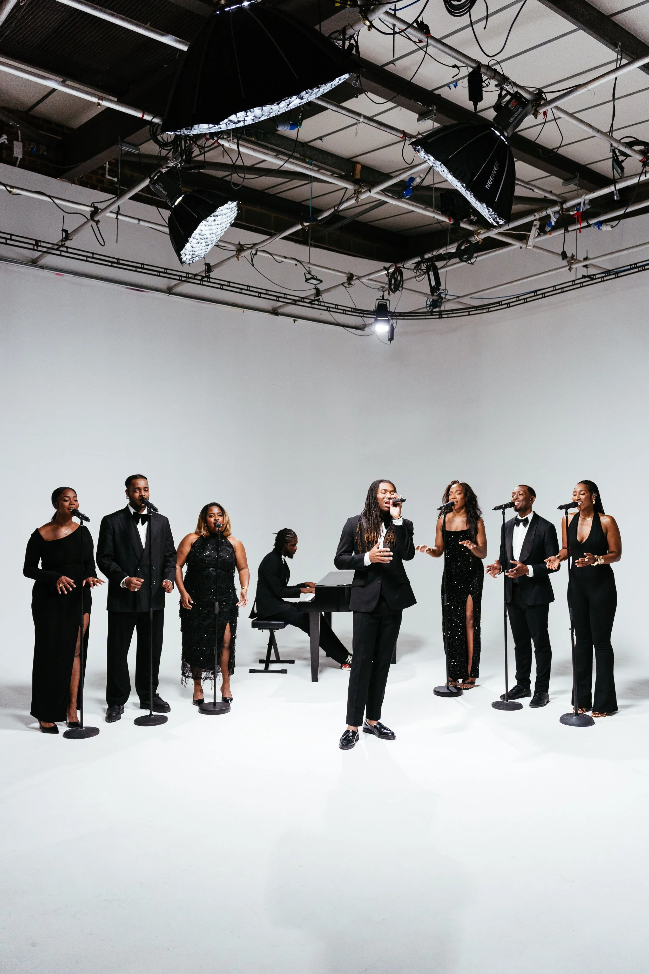 A diverse musical group performing in a studio with professional lighting, including a pianist, several vocalists in formal black attire, and a lead singer in the center.