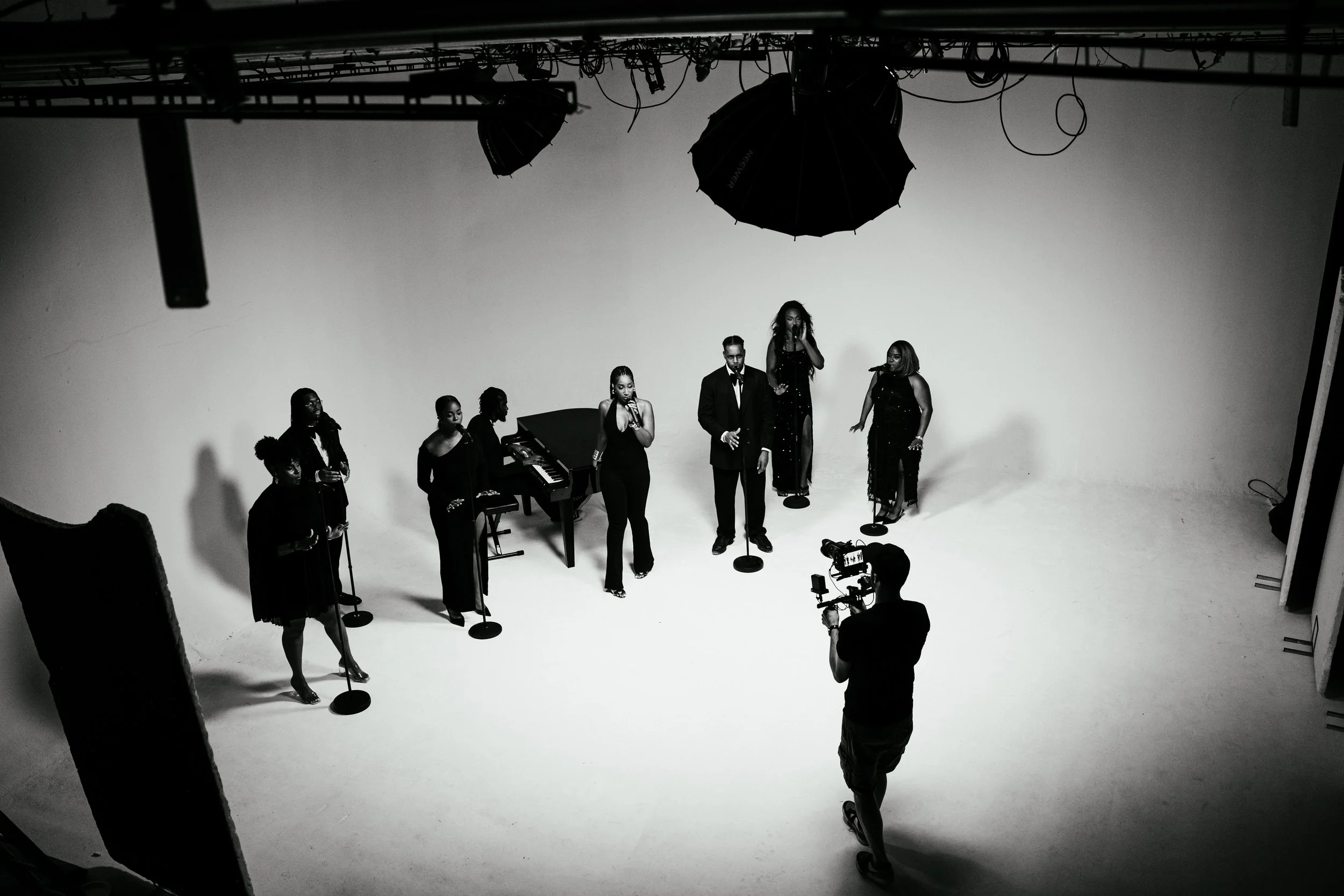 Black and white photo of six singers and one pianist performing in a studio. The singers are dressed in black and stand behind microphones, with a grand piano in the background. A videographer is filming the performance.