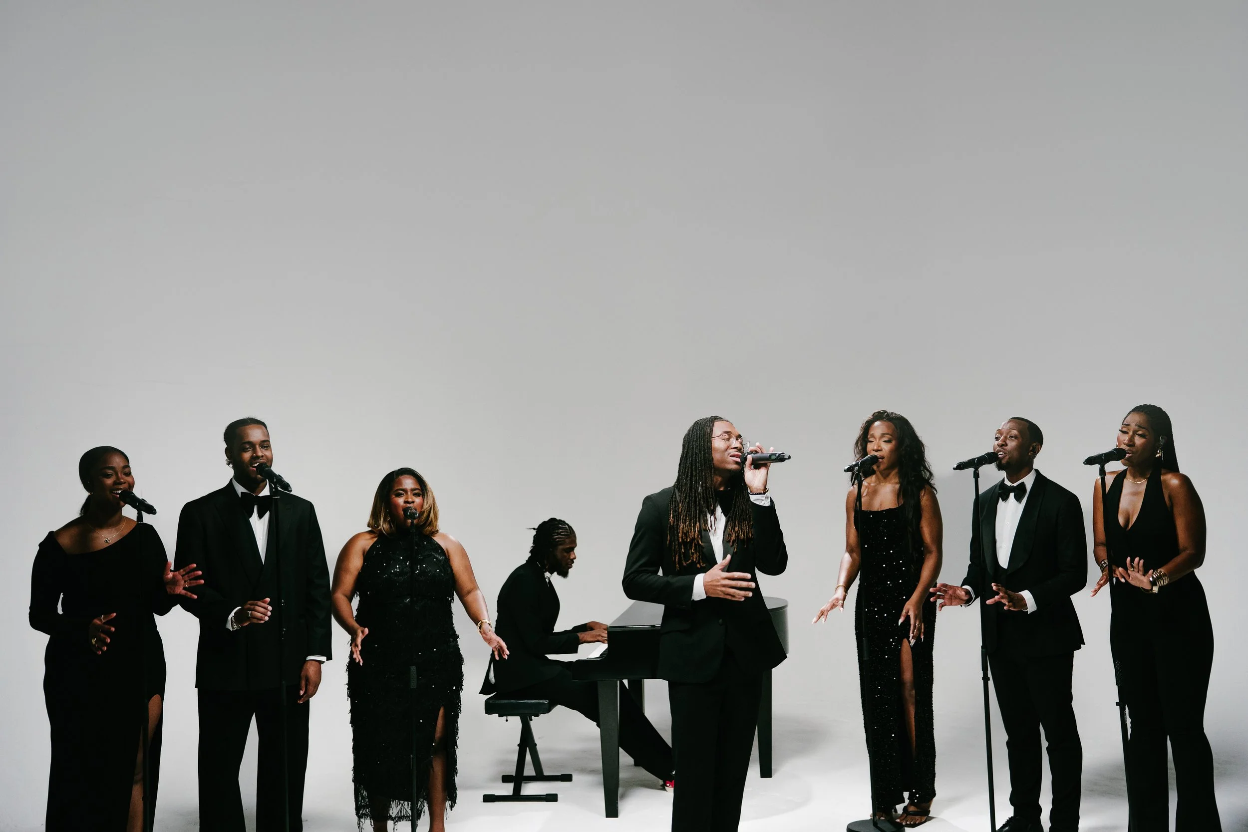 A group of eight singers dressed in formal black attire performing with microphones, accompanied by a pianist playing on a grand piano against a plain white background.