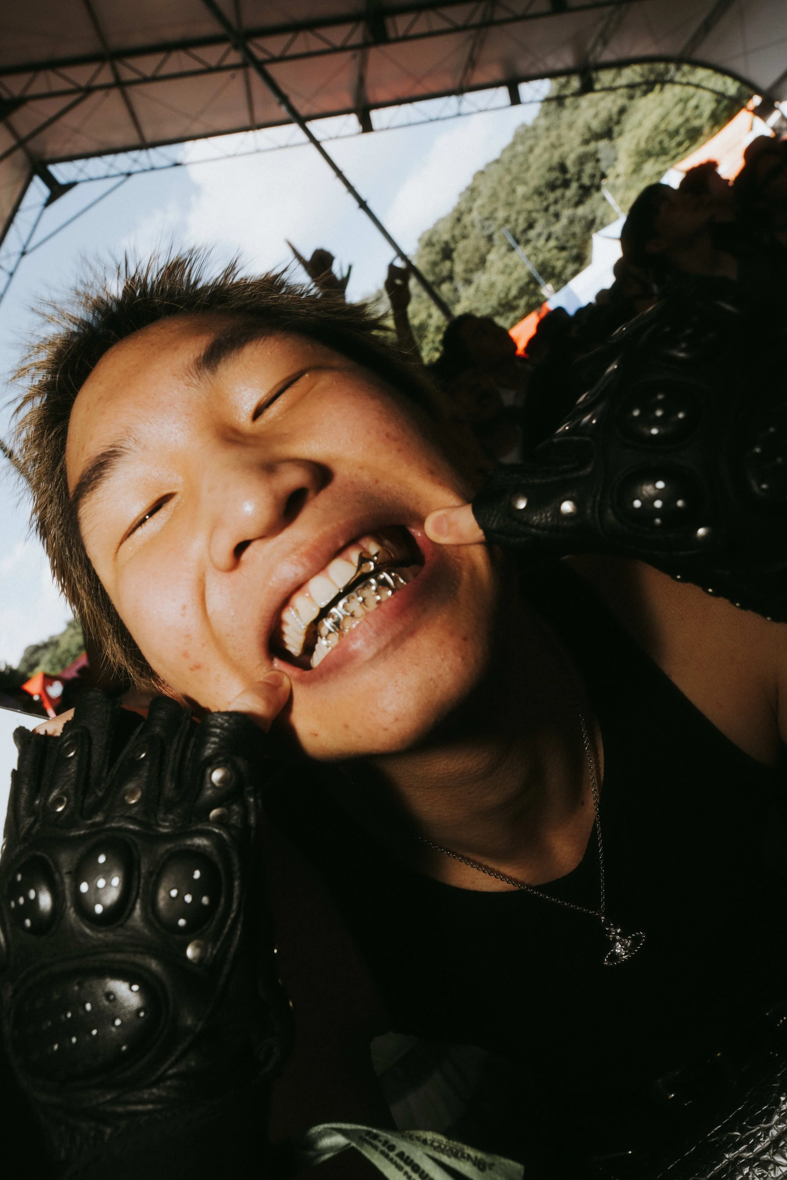 Person smiling broadly, showing gold teeth, with earrings and wearing black leather gloves and clothing, at an outdoor event or festival under a canopy.