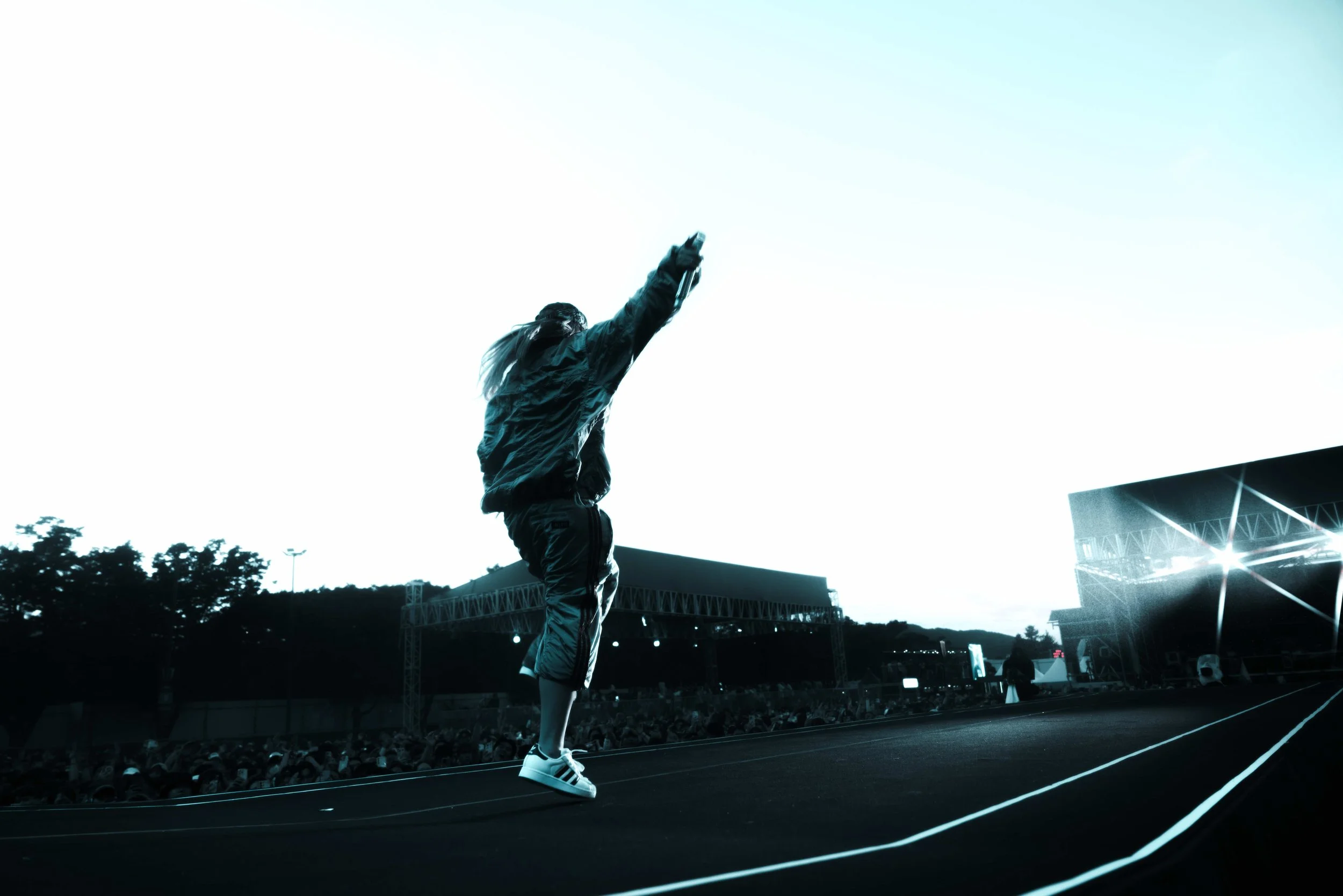 Person on stage at concert, with a crowd in the background, raising one arm, during an outdoor event in the evening.