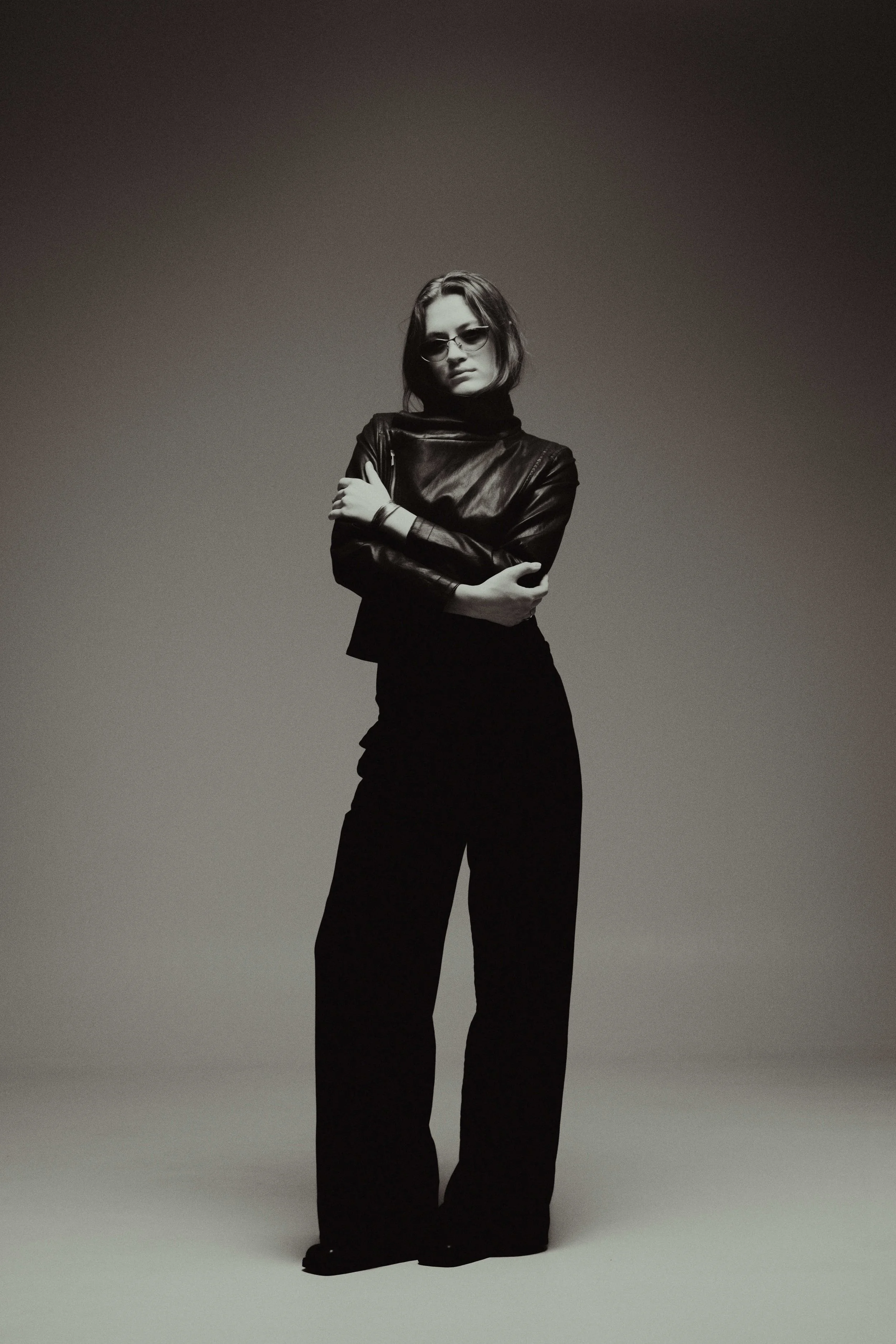 A woman with sunglasses, wearing a black leather jacket and black wide-leg pants, stands with her arms crossed against a gradient background.
