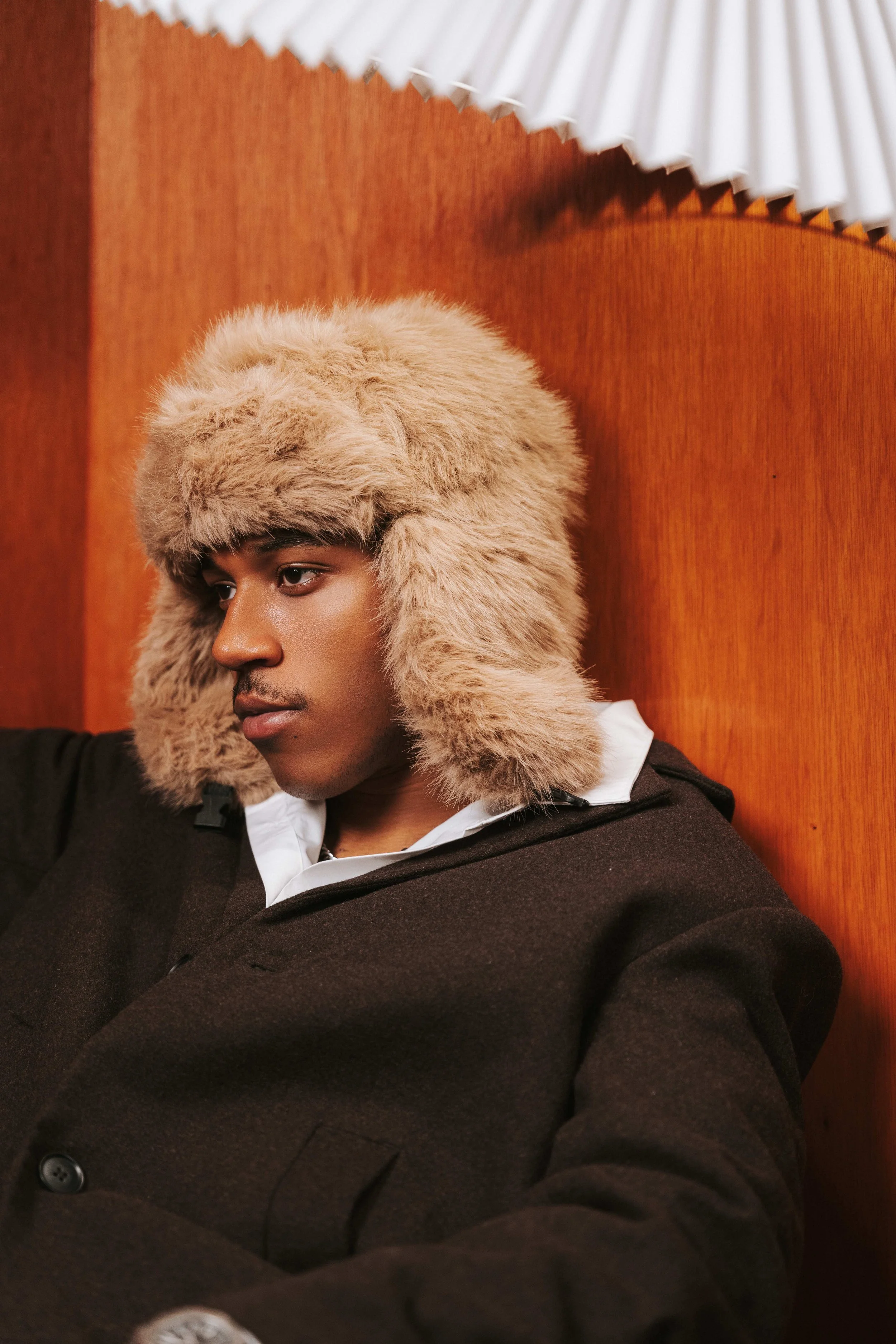 Young man sitting against a wooden wall, wearing a furry beige winter hat and a dark coat.