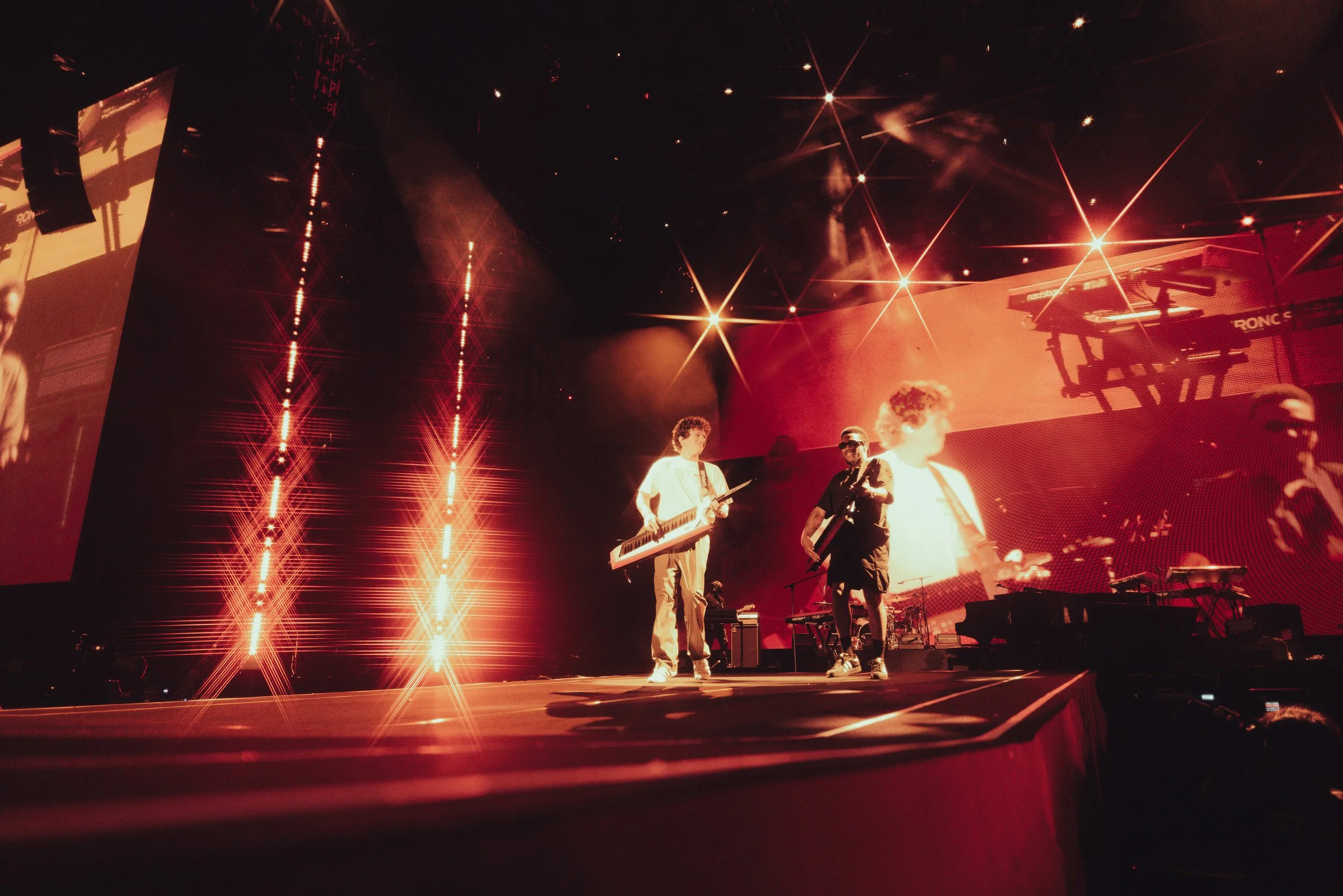 Three musicians on stage performing with large red screens and bright stage lights in the background. One is playing a keytar and the other two are holding guitars.