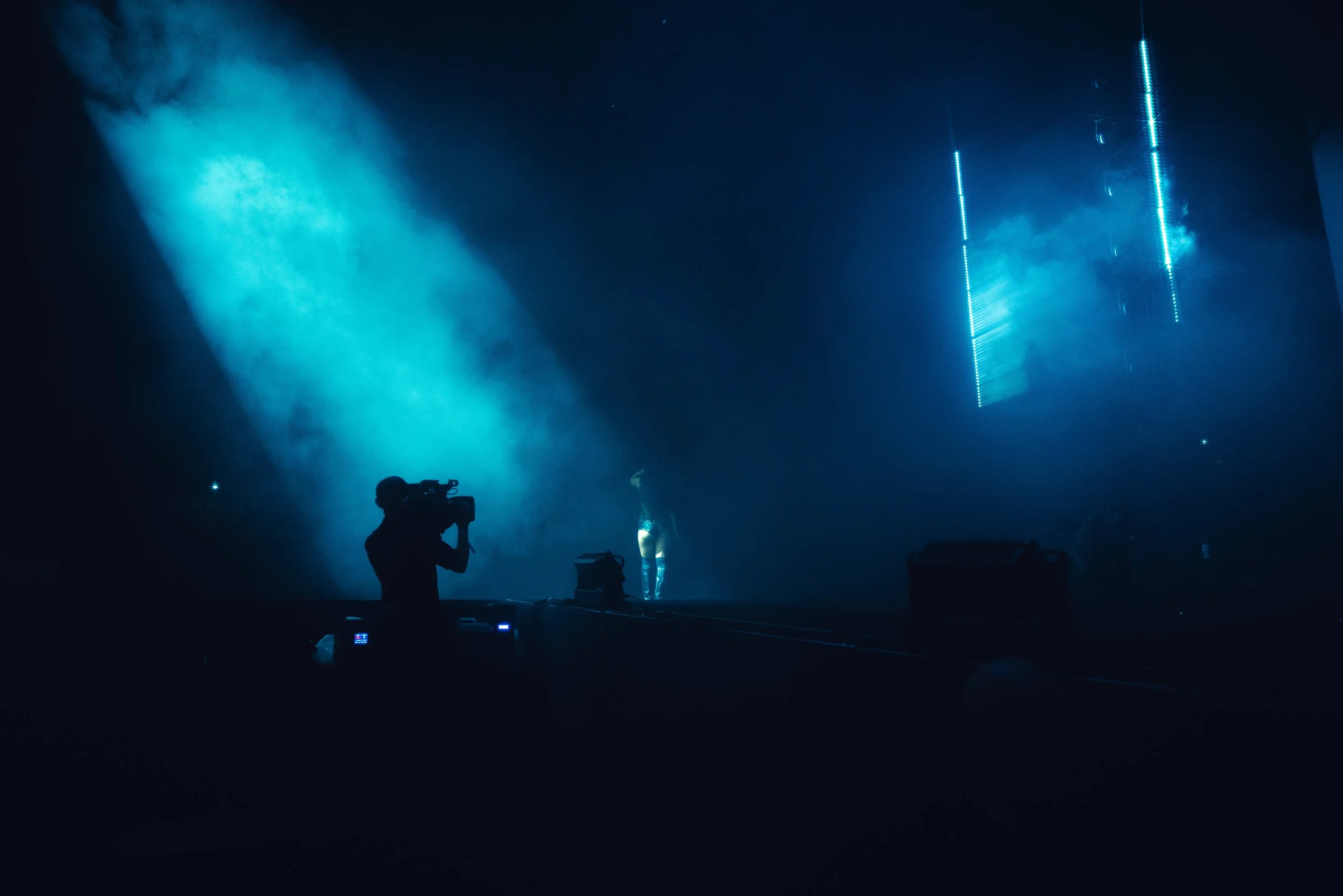 A silhouette of a photographer holding a camera on a studio stage with blue lighting and fog, and a partially visible performer in the background.