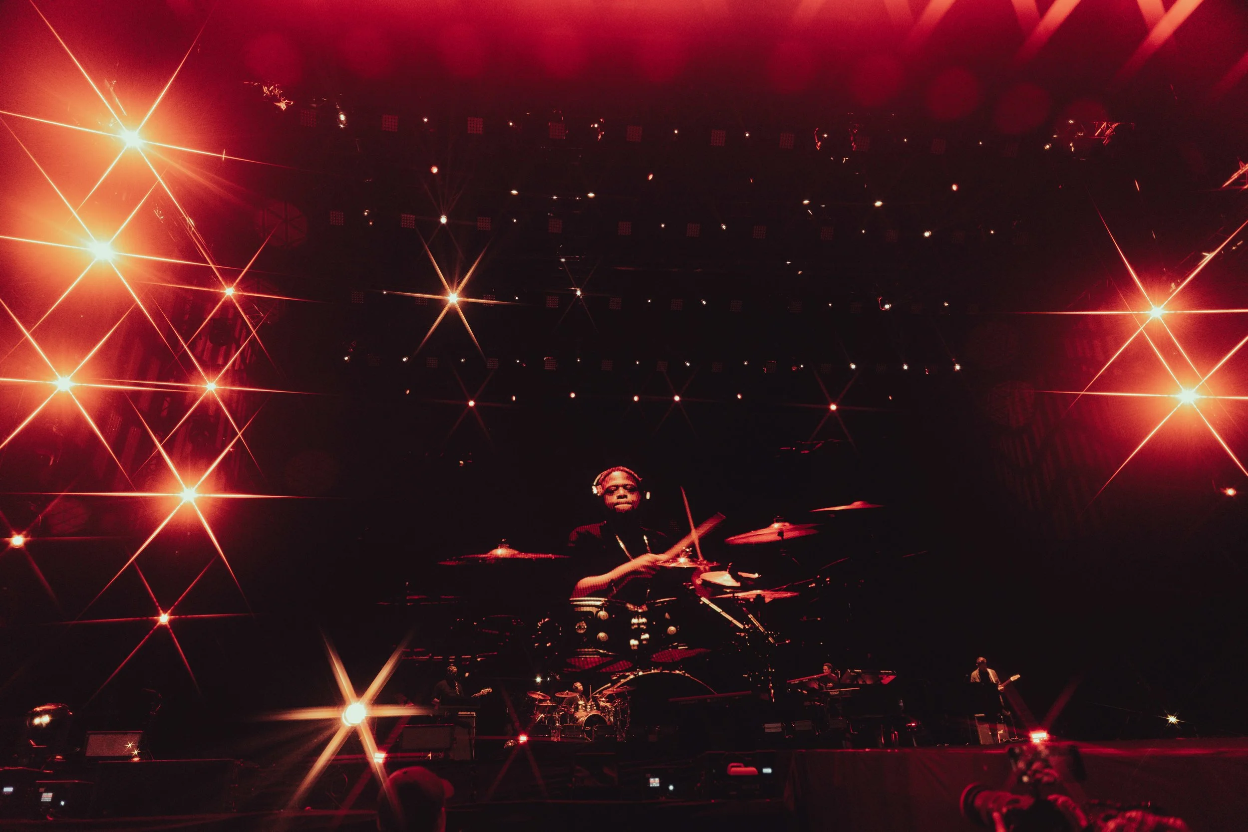 A musician playing drums on stage with large red and orange lights and starburst effects in the background.