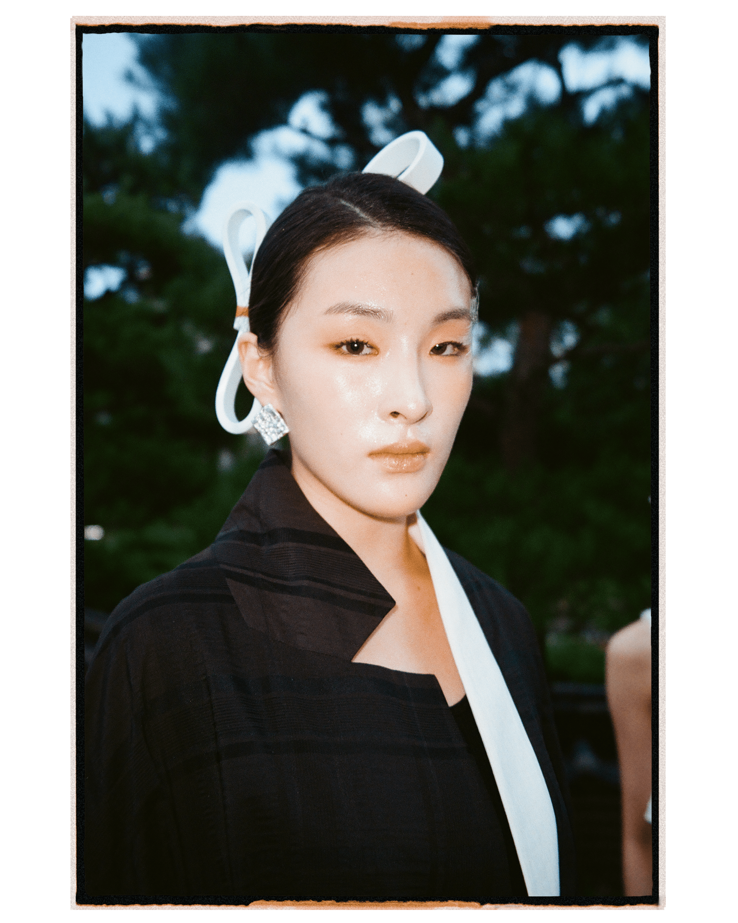 A portrait of a woman with slicked-back dark hair, wearing a large white sculptural hair accessory, diamond earrings, and a black jacket with a white lapel, outdoors with blurred trees in the background.