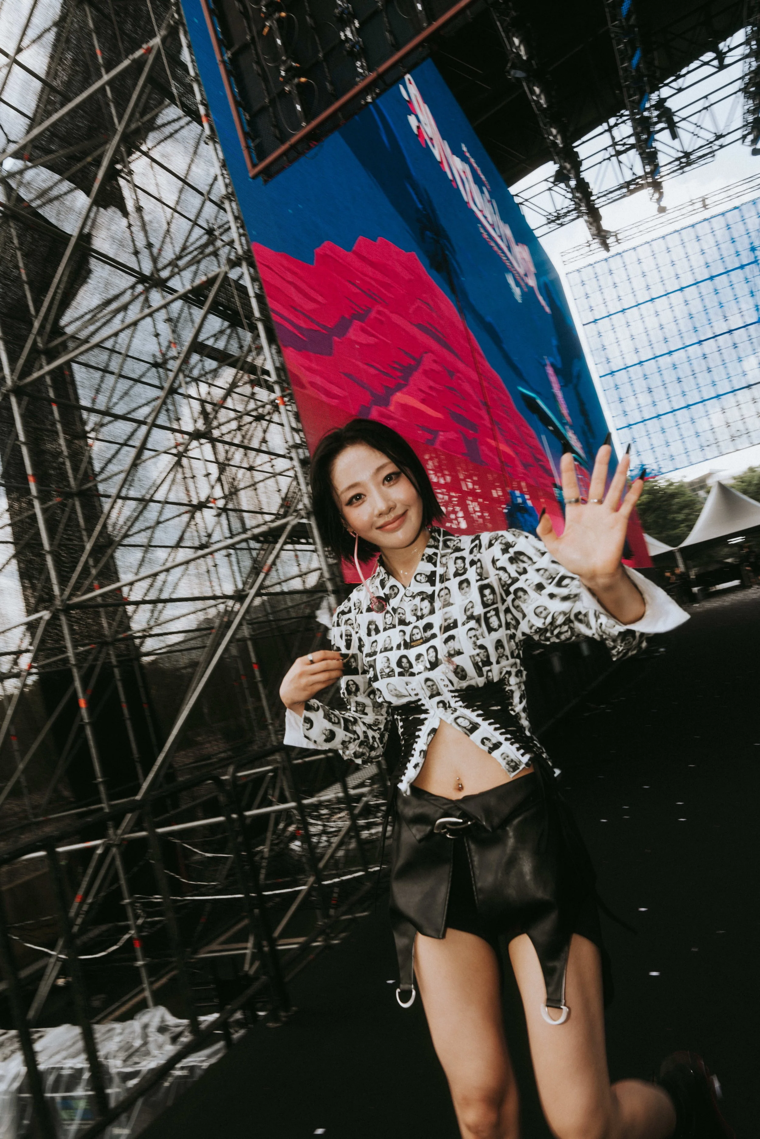 A young woman waving at the camera during an outdoor event or concert, with metal scaffolding and a large digital screen in the background.