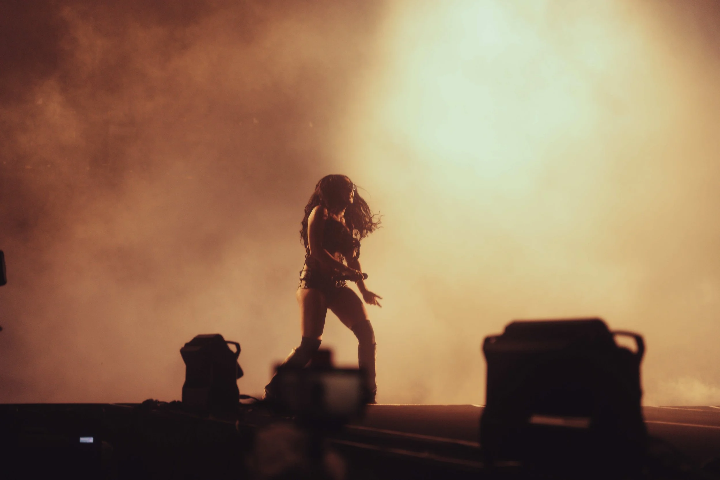 A person with long hair dancing on stage with bright yellow and orange lighting in the background, surrounded by stage equipment.