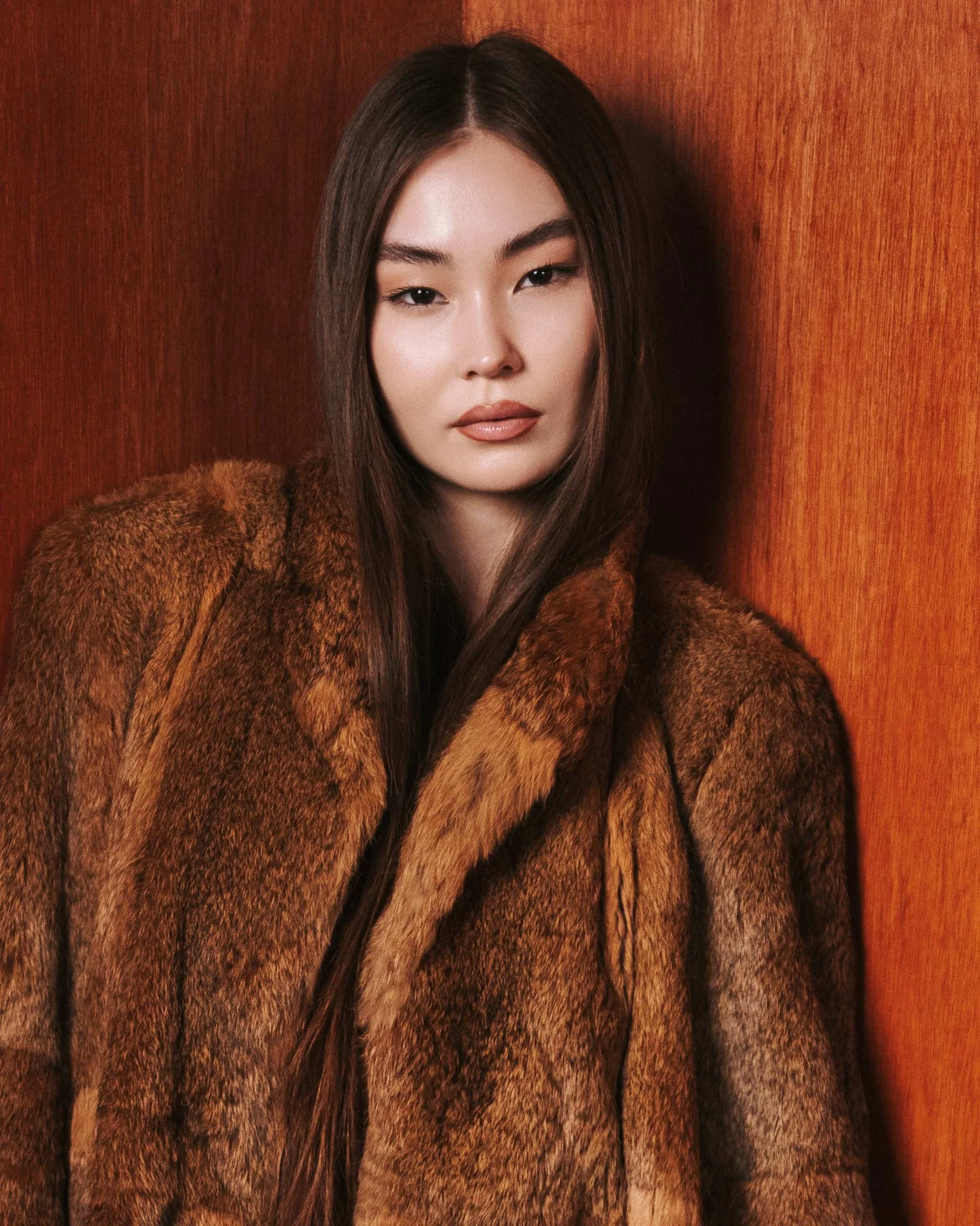 A woman with long dark hair wearing a brown fur coat, standing against a wooden wall.