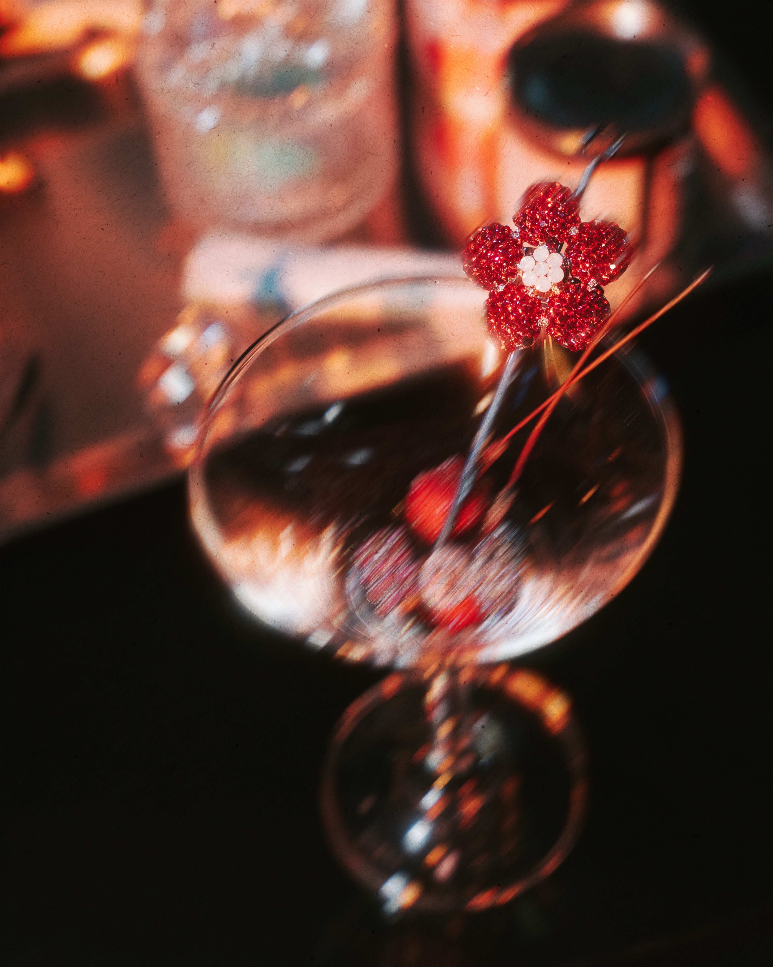 A close-up of a wine glass with red berry garnish and a flower-shaped decoration. The background features a blurred bottle and other items.