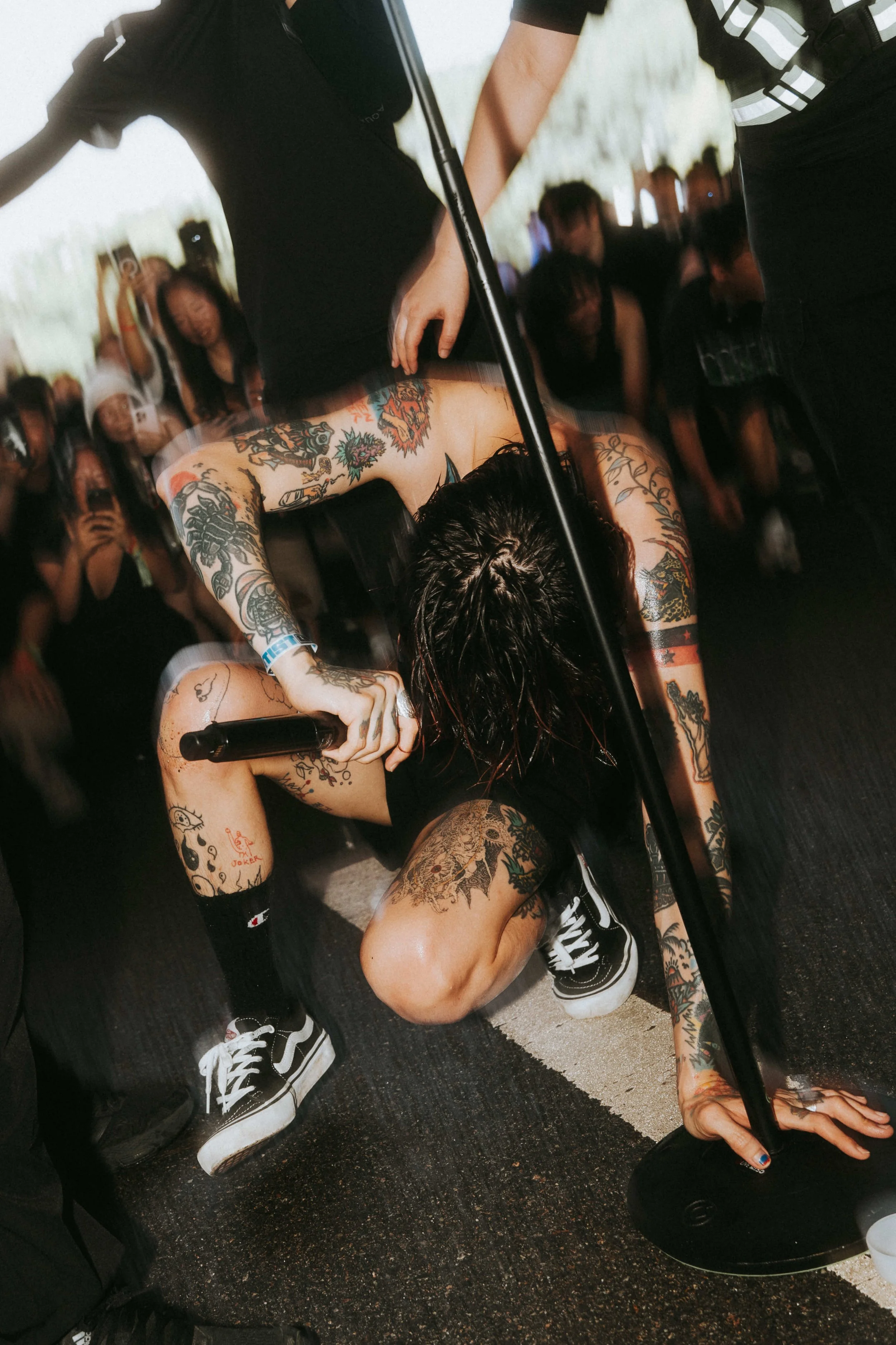 A tattooed performer crouching on the floor holding a microphone, surrounded by an audience of people taking photos and watching.