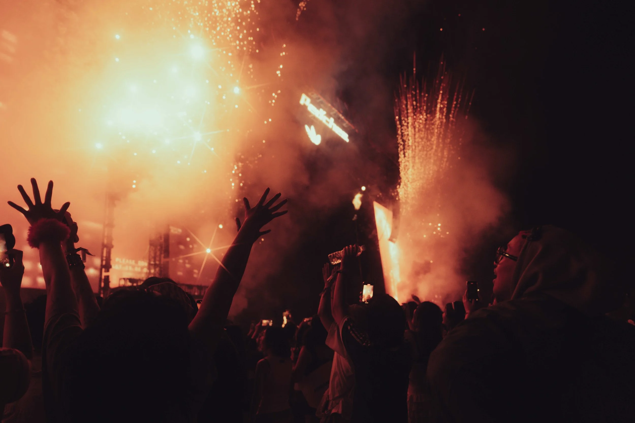 Crowd watching a fireworks display at night with raised hands and smartphones capturing the moment.