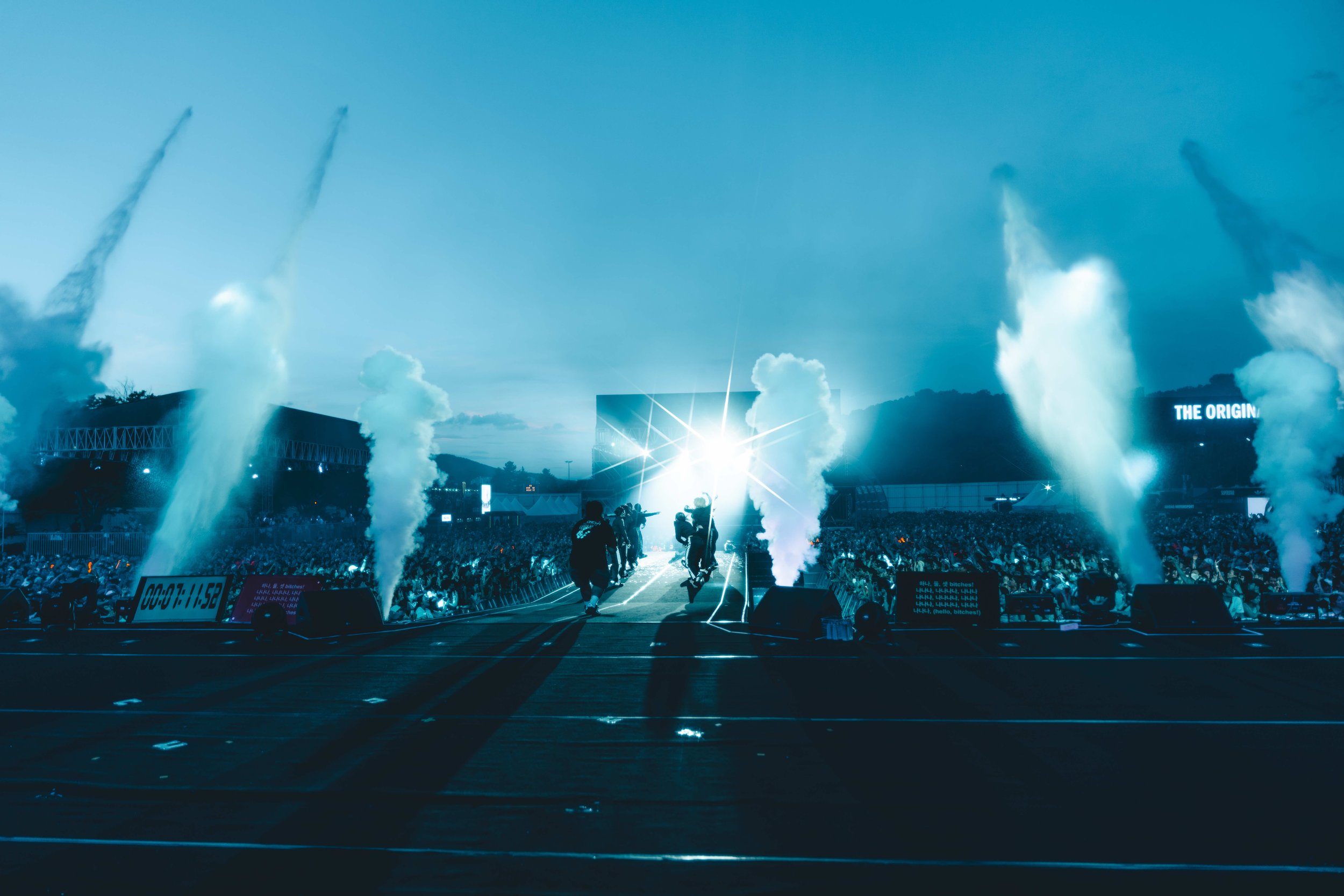 A concert or festival stage with bright lights, smoke effects, and a large crowd, taken during the evening or twilight.