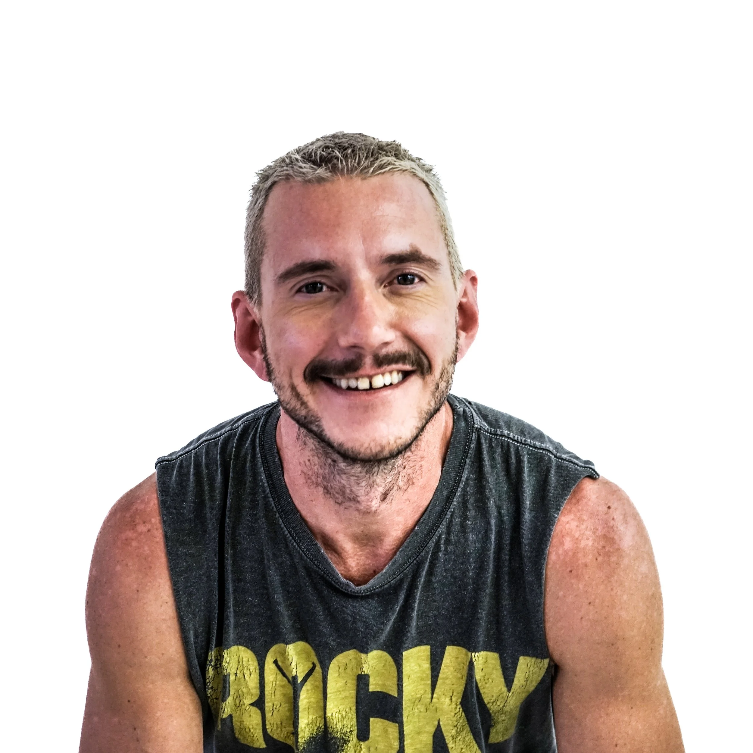A smiling man with short blonde hair, a beard, wearing a sleeveless black T-shirt with yellow text, against a plain white background.