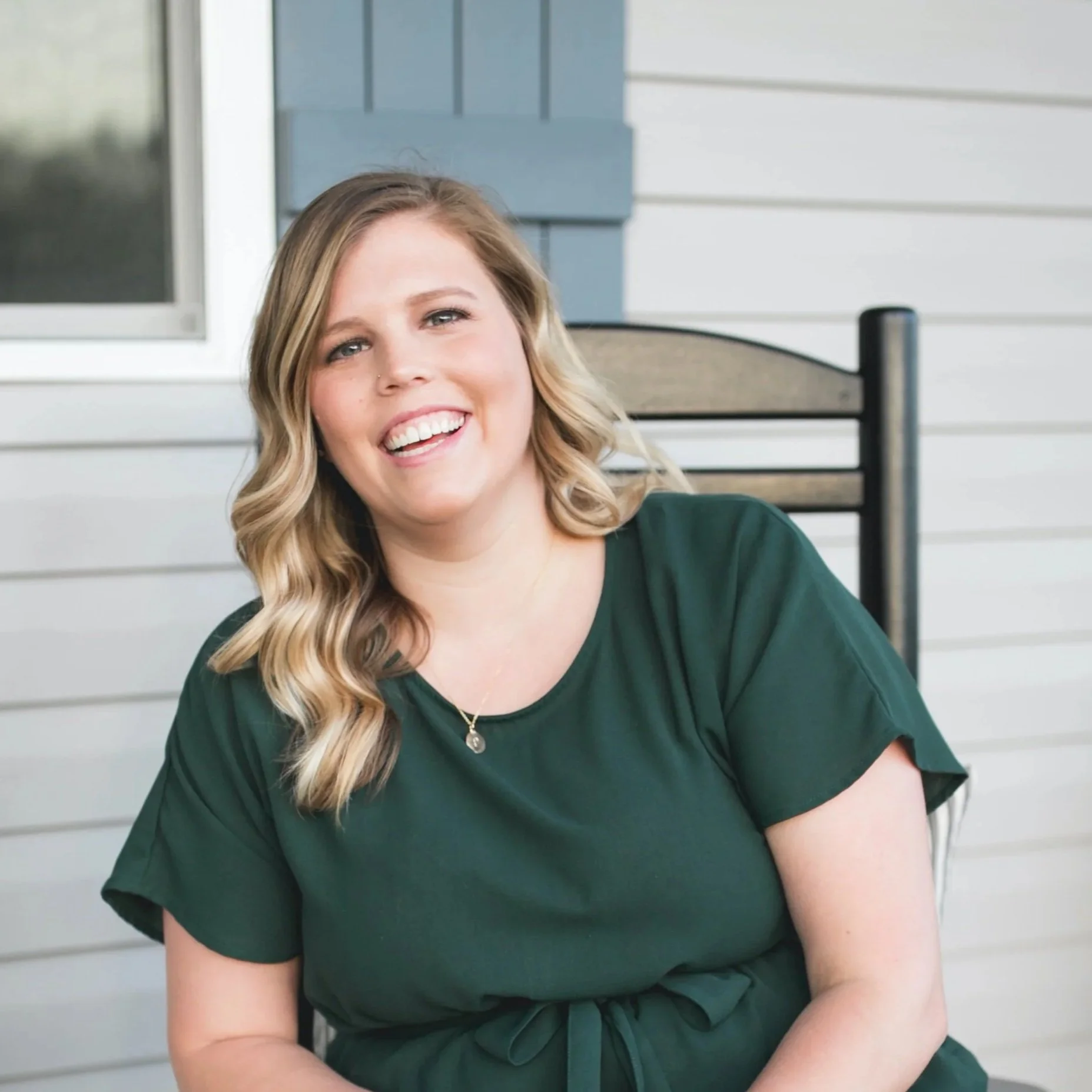 A woman with blonde hair wearing a dark green dress is sitting on a black rocking chair on a porch, smiling at the camera. The background features light-colored siding and a small plant.