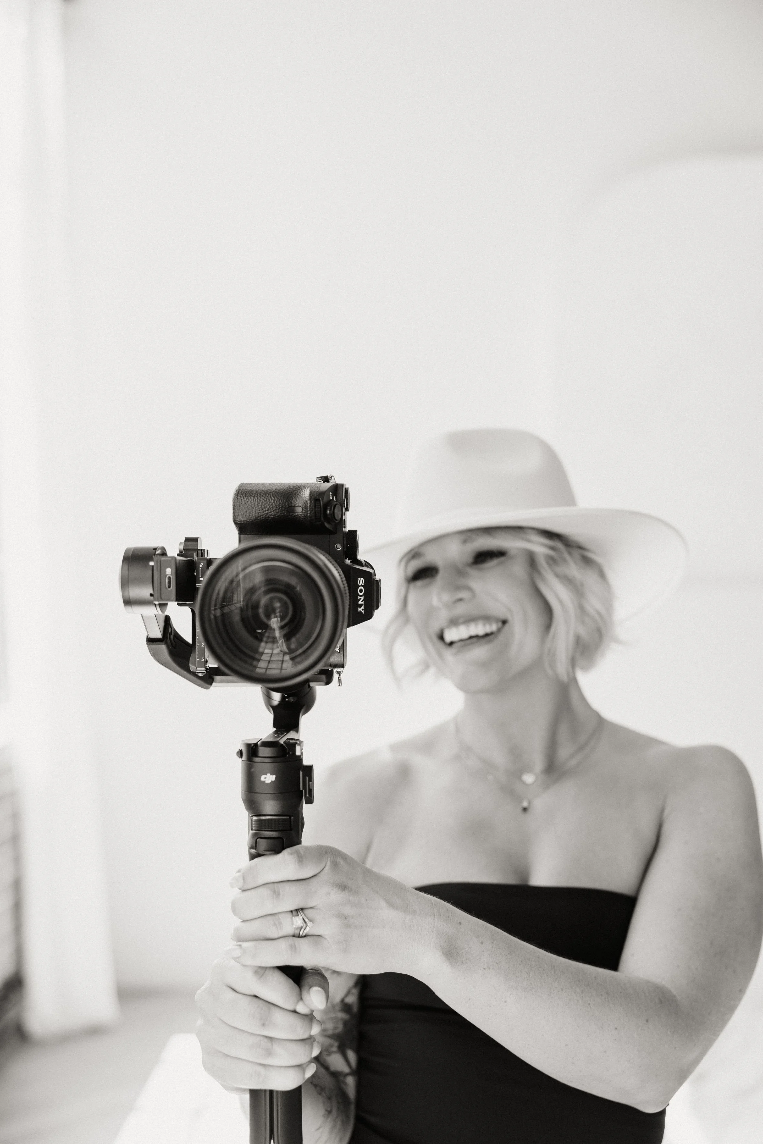 A woman with blonde hair wearing a wide-brimmed hat and strapless top taking a selfie with a camera on a tripod.