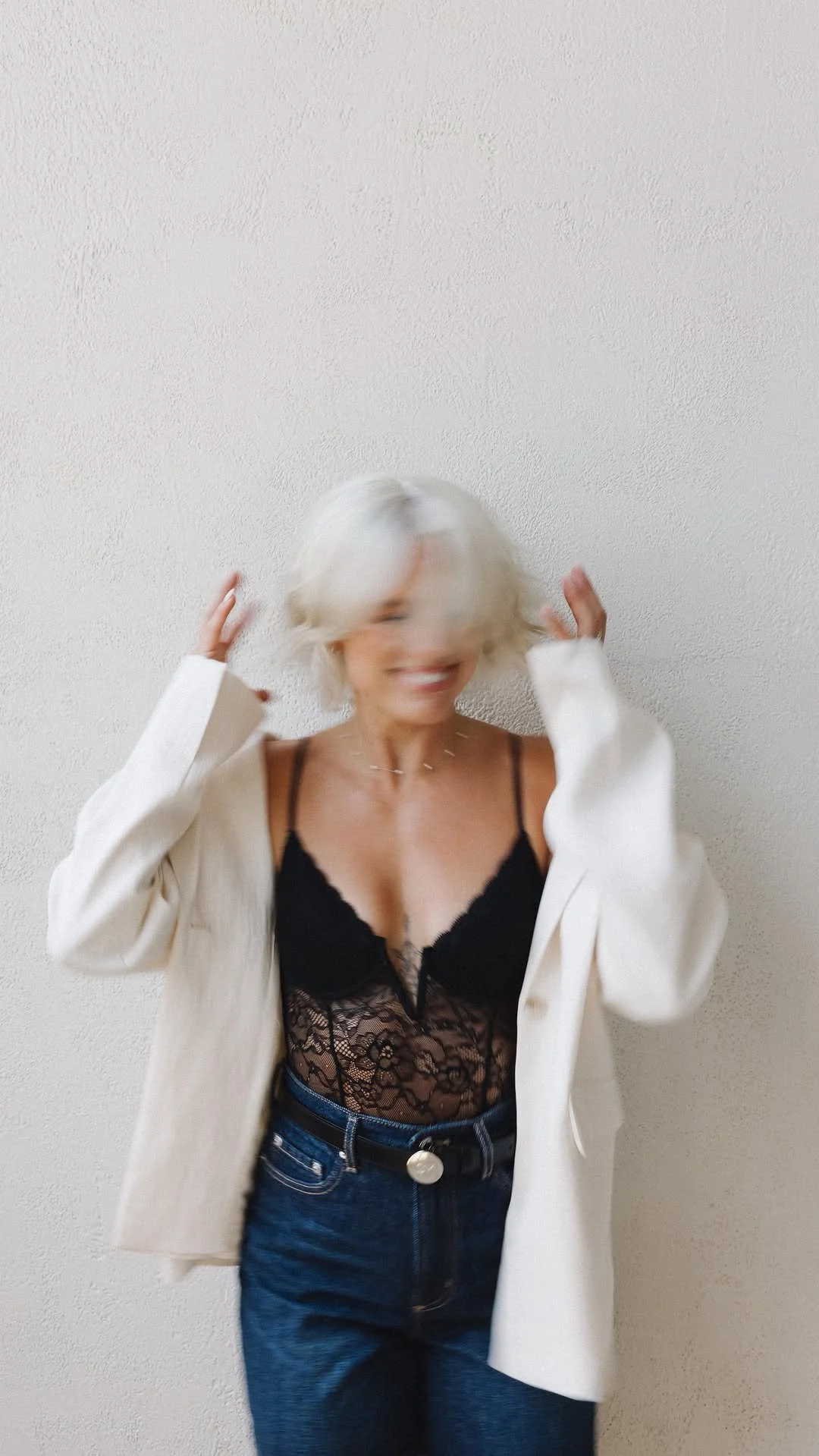 A woman with short white hair, smiling, wearing a black lace top, a beige blazer, and blue jeans, standing against a white textured wall.