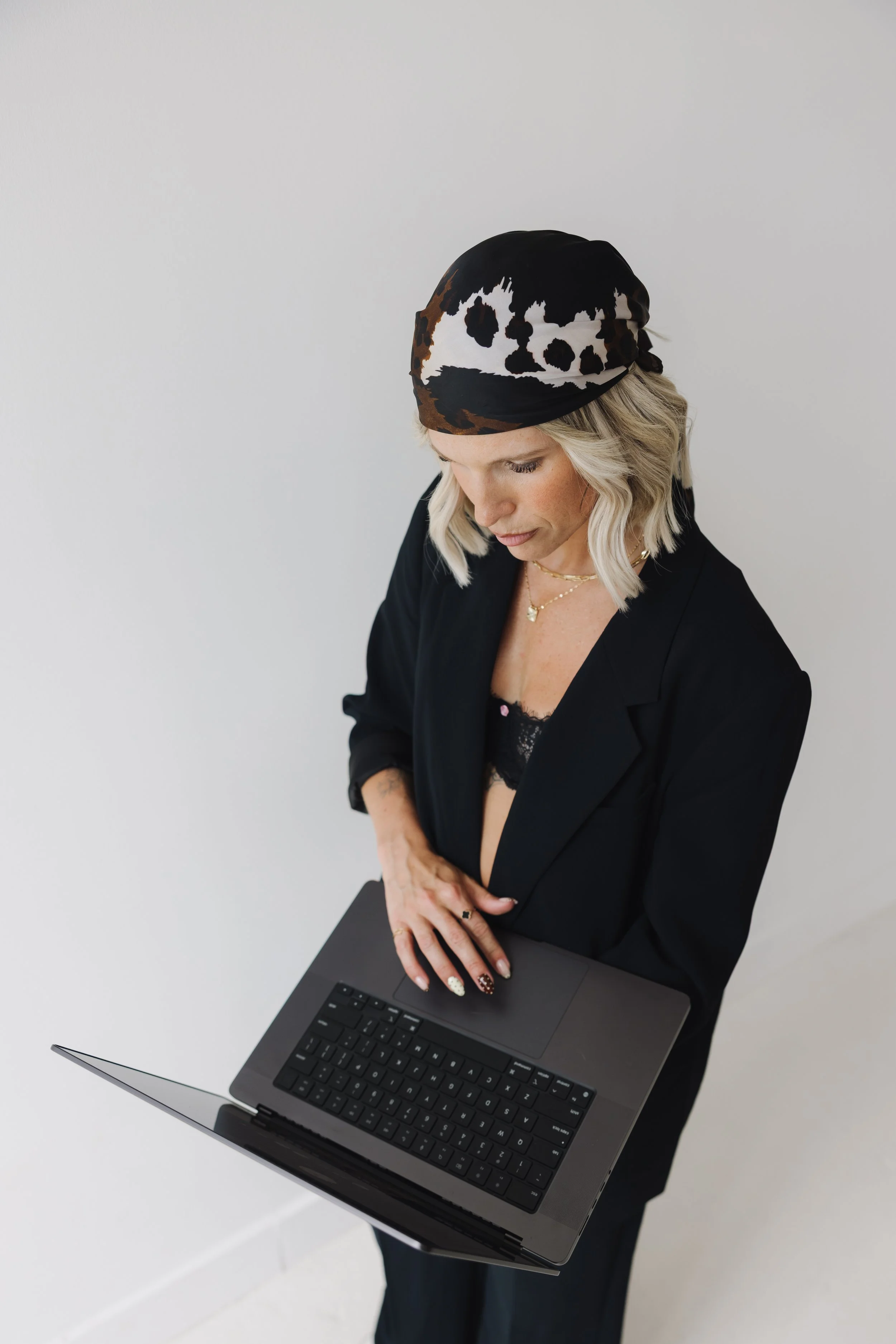 A woman with blonde hair wearing a black blazer, lace top, and a black, white, and brown patterned headscarf, using a laptop on a white table against a plain, light-colored wall.