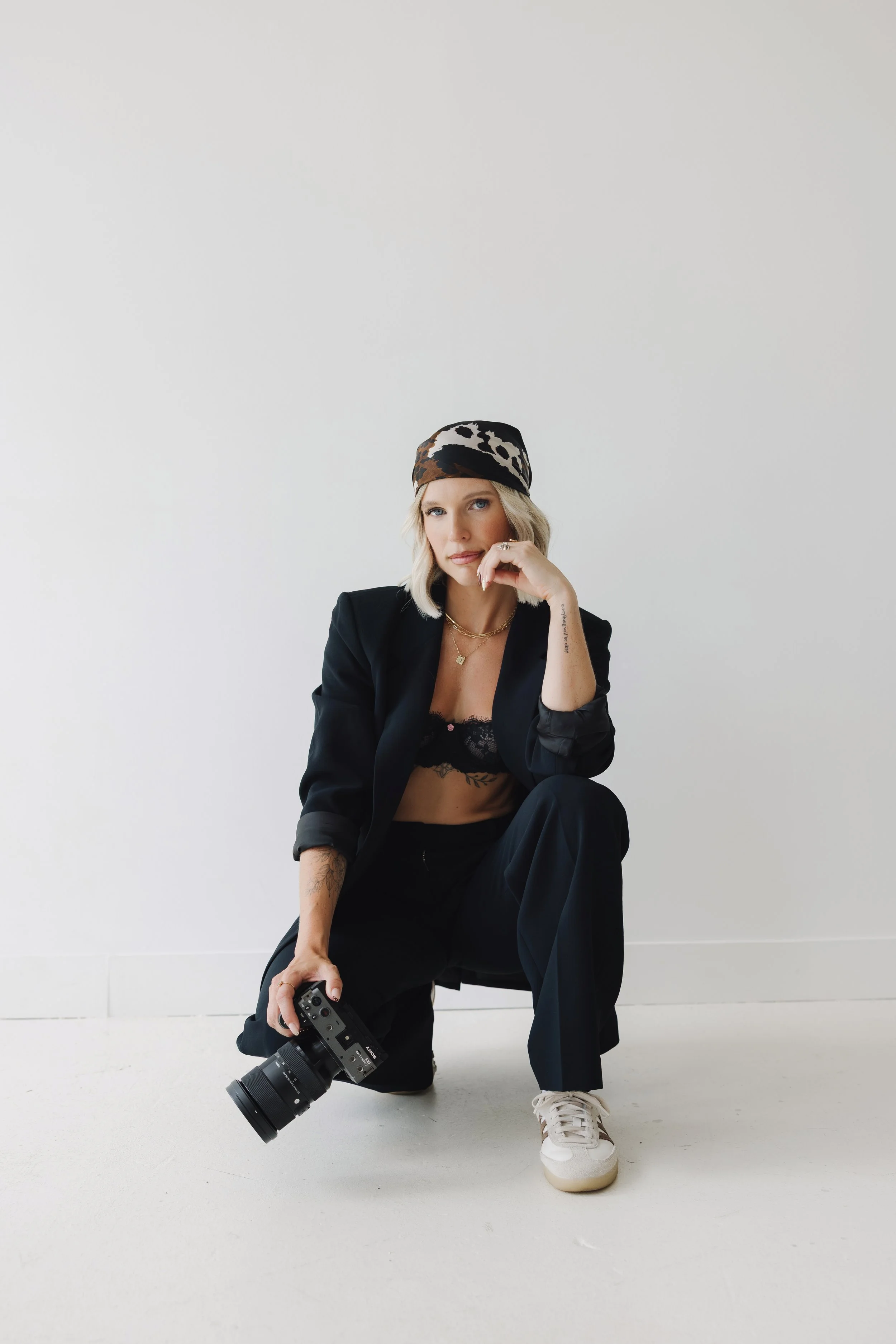 A woman with blonde hair wearing a leopard-print headscarf, a black blazer, lace black top, and black pants, holding a camera and sitting on the floor against a plain light gray background.