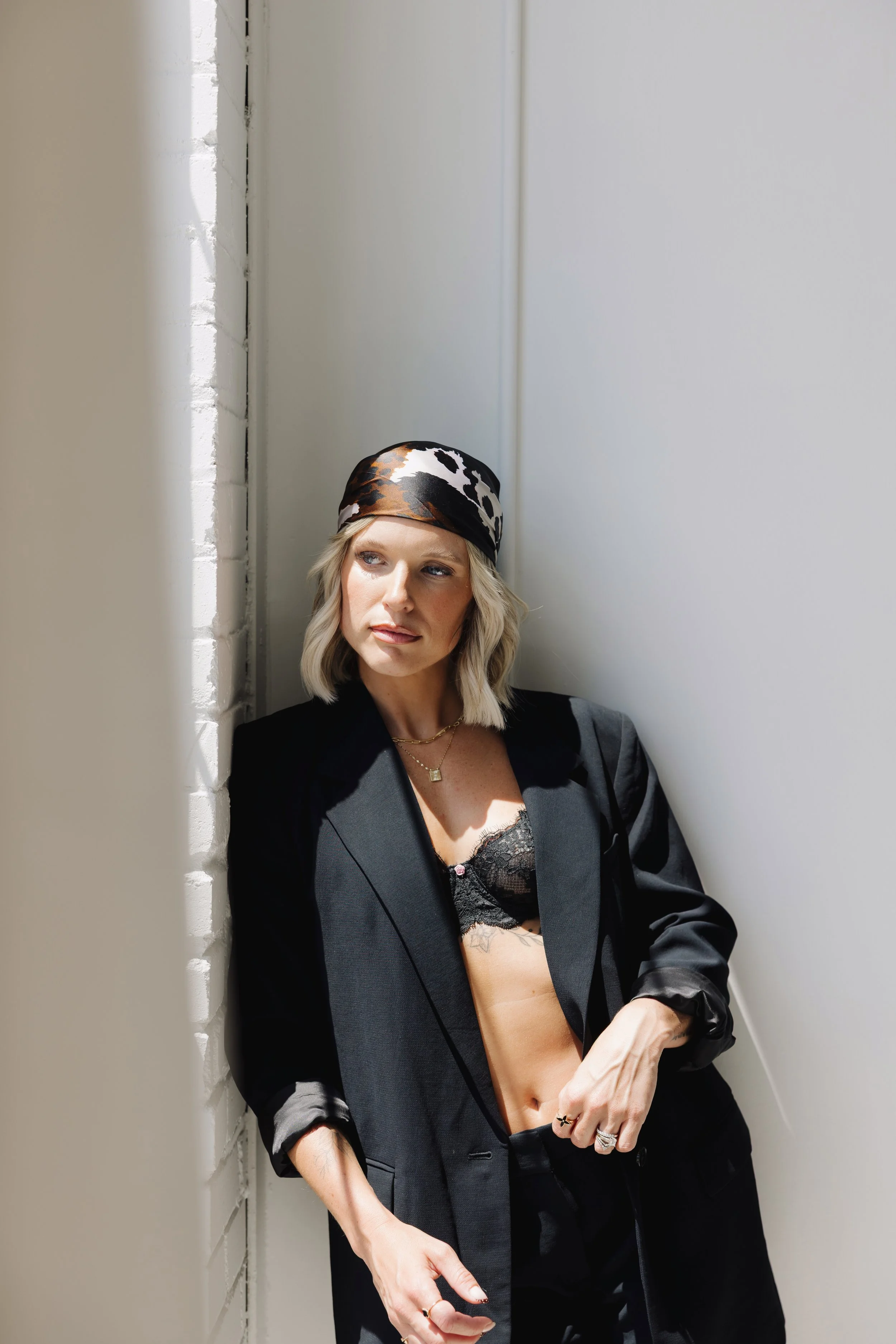 A woman with blonde hair slicked back, wearing a patterned headscarf, a black blazer, and a black lace bralette, poses against a white brick and painted wall with sunlight highlighting her face and upper body.