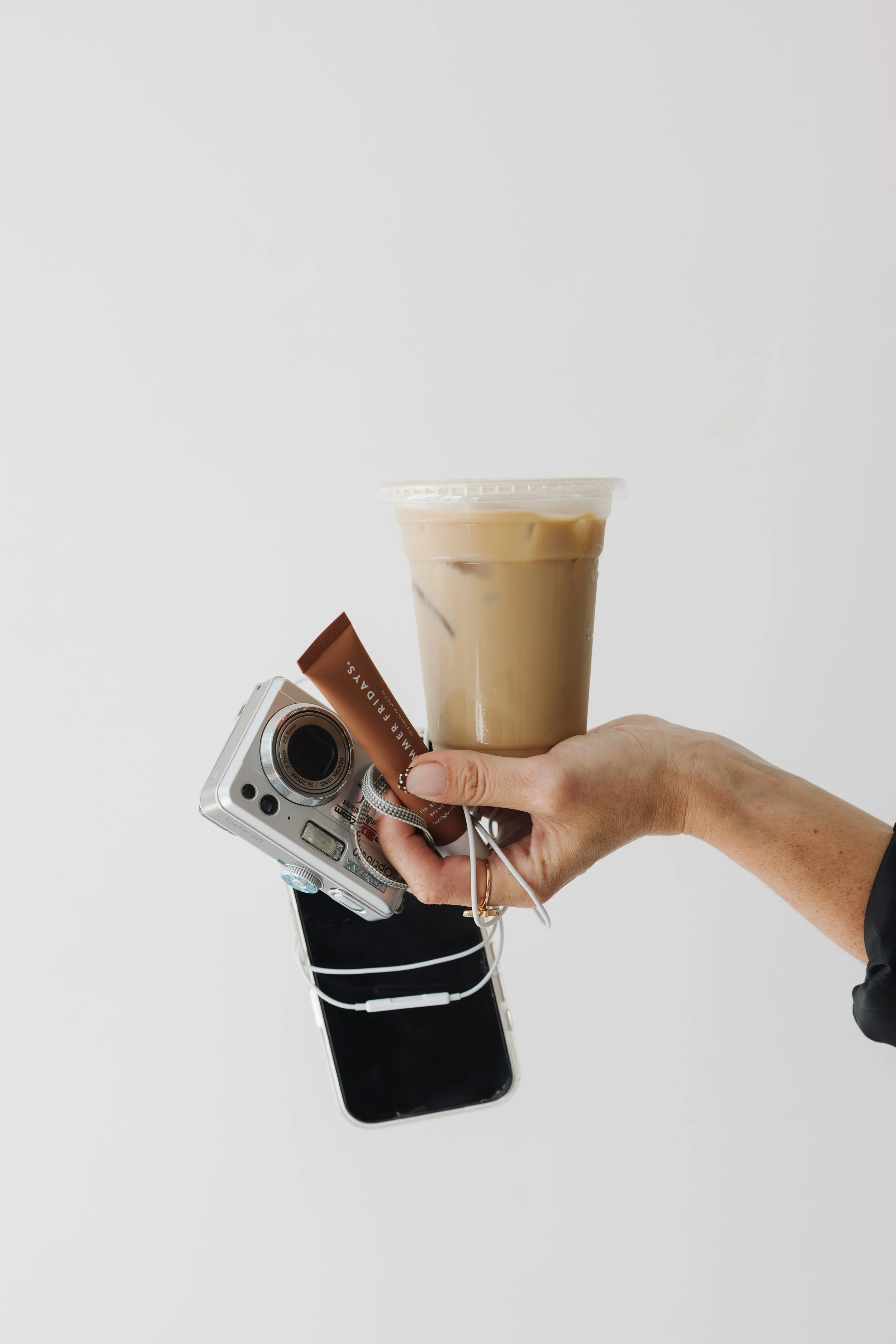 Hand holding a smartphone with a camera, a tube of skincare product, a latte in a clear plastic cup, and earphones against a white background.