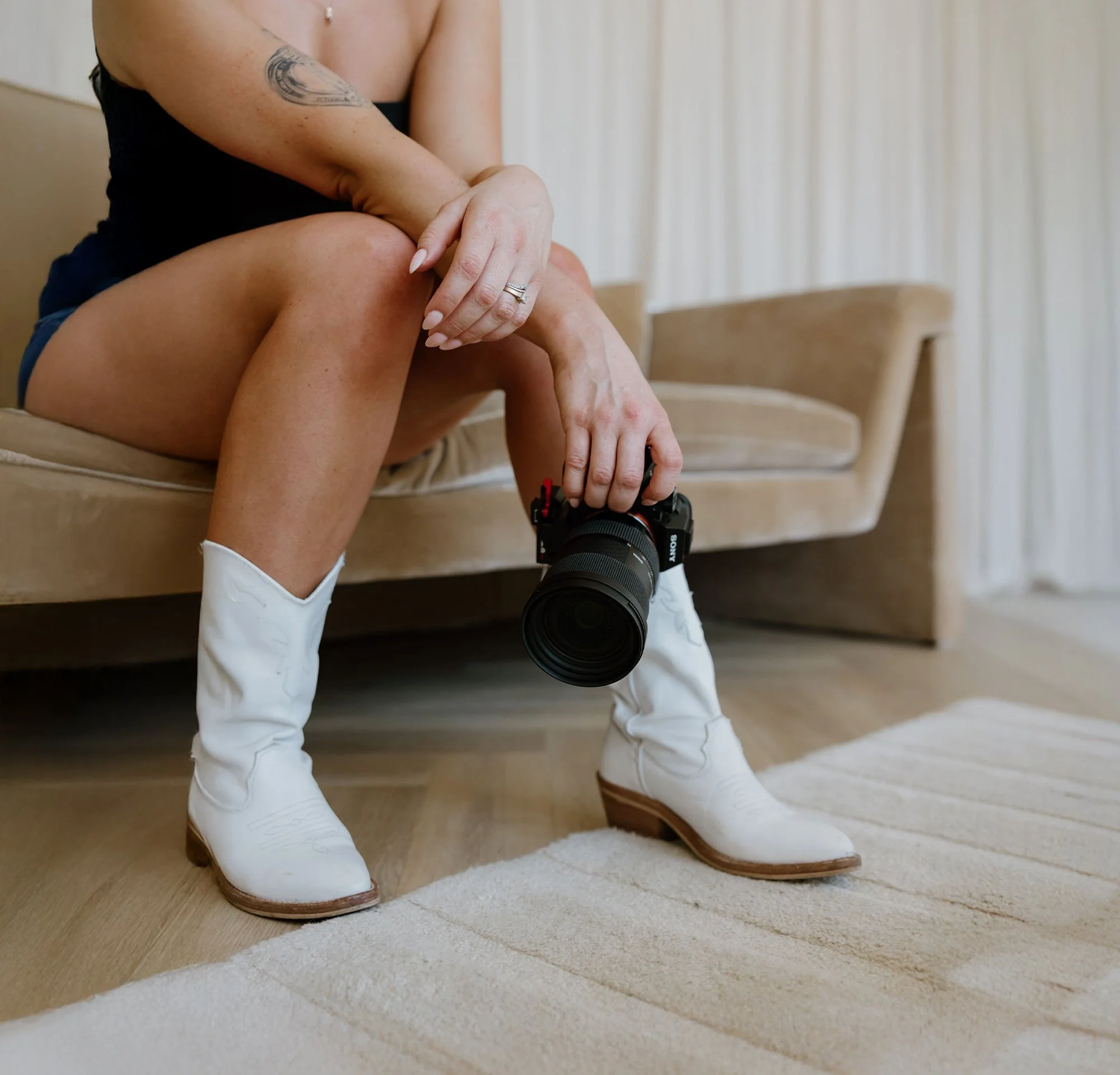 Person sitting on a beige couch, holding a camera pointed downward, wearing white cowboy boots and shorts, in a room with wooden flooring and cream curtains.