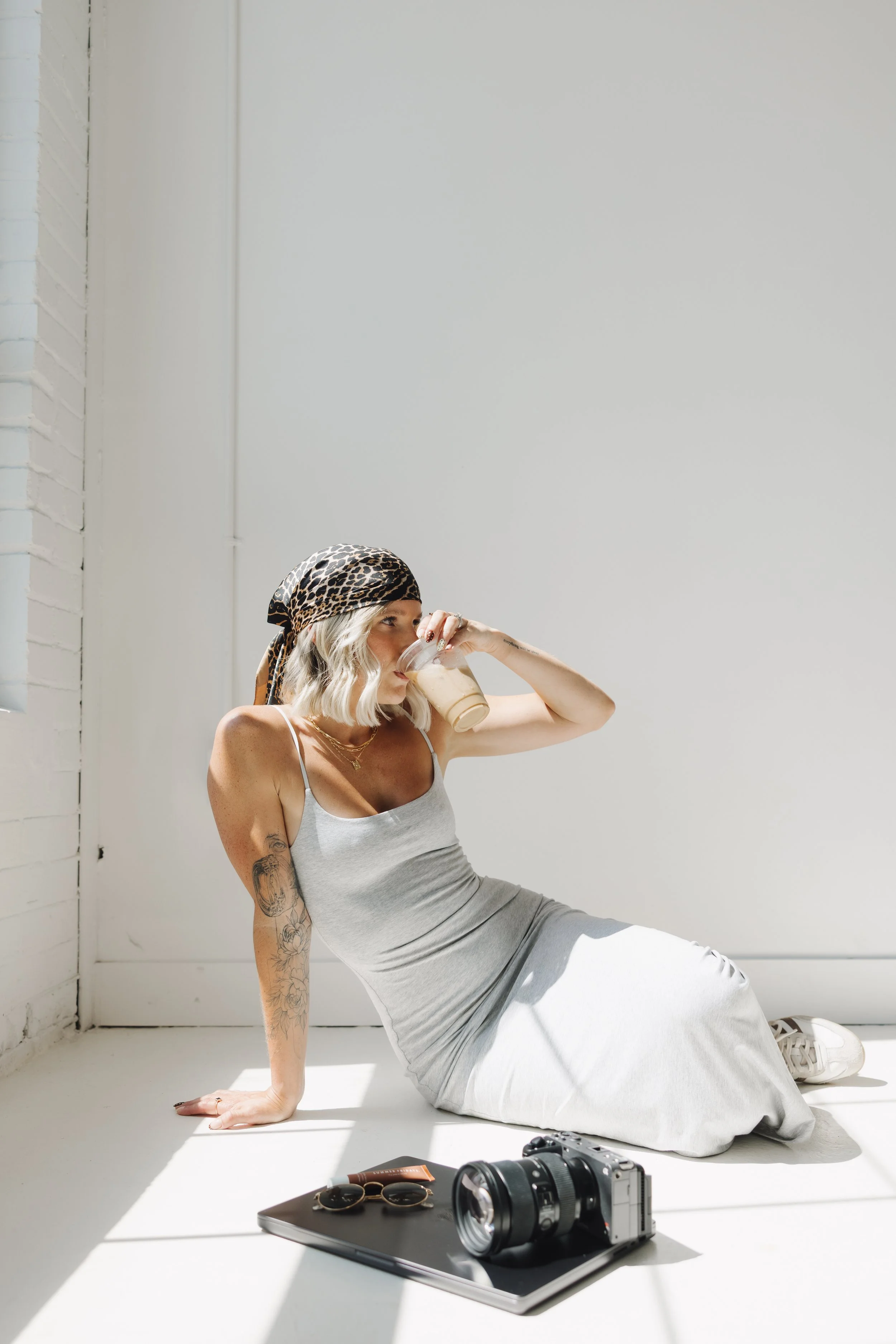 Woman with tattoos and a leopard print headscarf sitting on the floor, drinking from a glass of iced coffee or latte, in a brightly lit room with white walls and a white brick section. A camera, sunglasses, and a notebook are on the table in front of her.