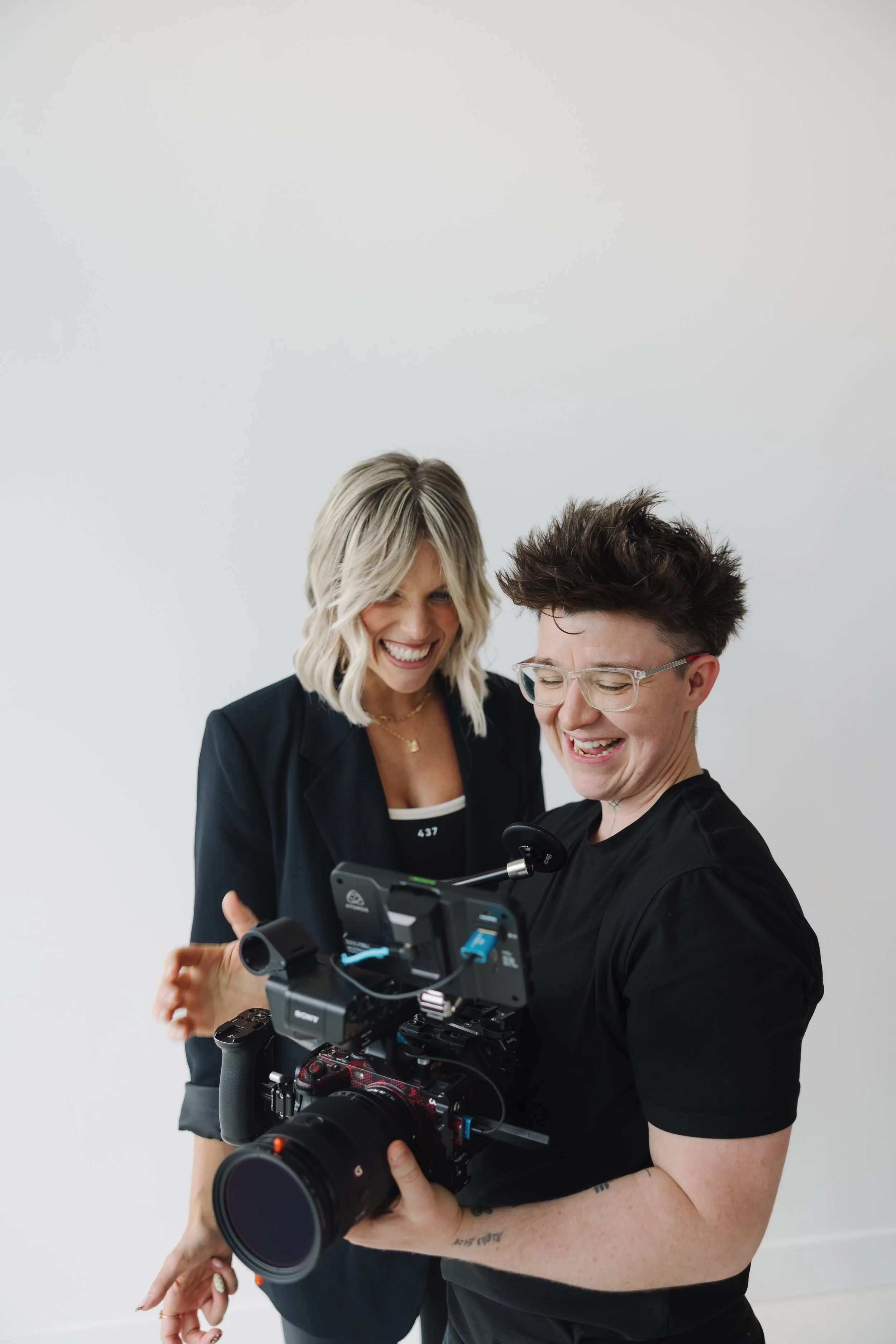 Two women are smiling and looking at a camera. The woman on the left has blonde hair, is wearing a black blazer, and is laughing. The woman on the right has short, dark, spiky hair and glasses, and is holding a professional camera. They are in front of a plain white wall.