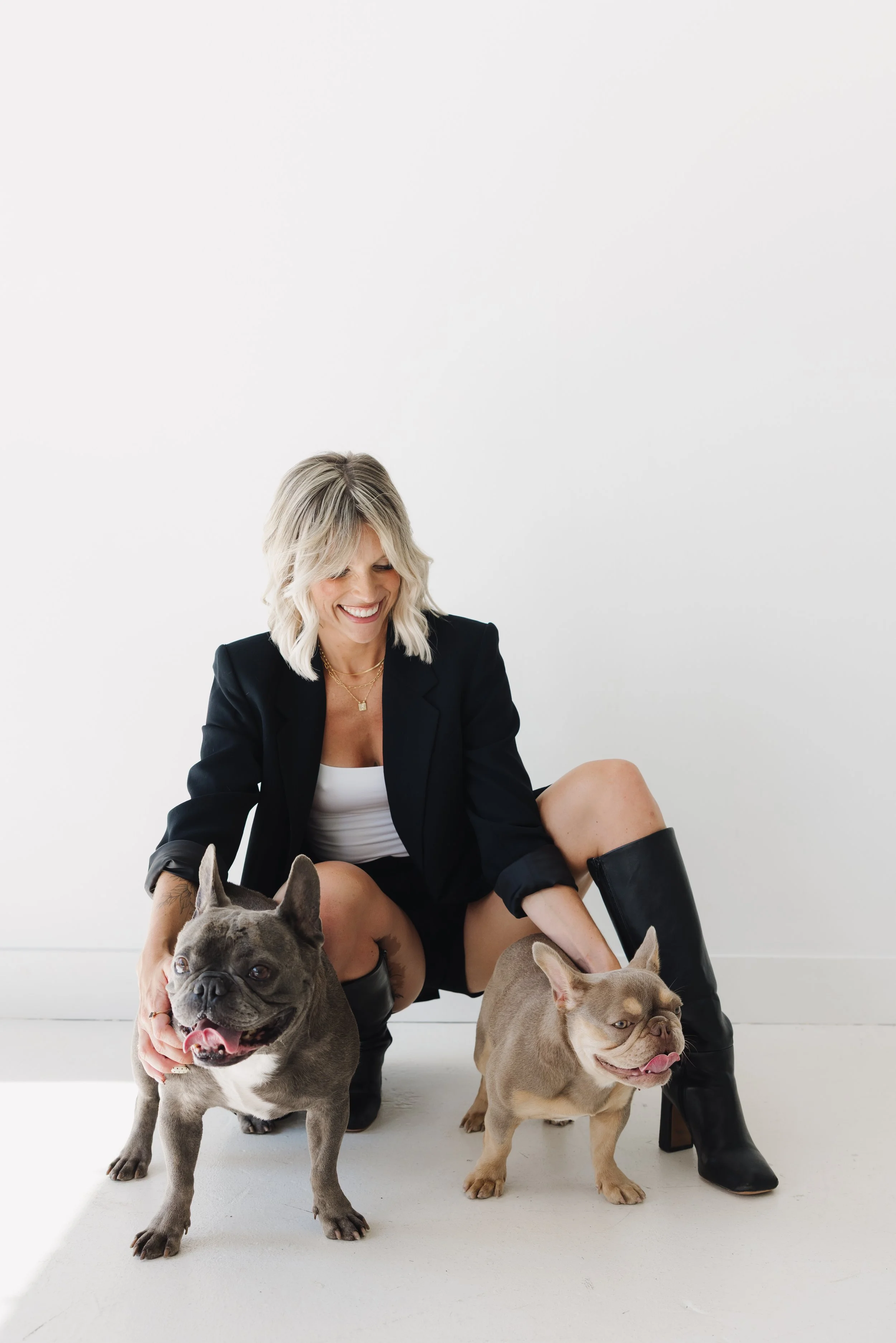 A woman with blonde hair smiling, dressed in a black blazer and shorts, is seated on the floor with two French Bulldogs. One bulldog is gray with a white chest and paws and is standing in front, while the other is light brown with a darker snout, sitting beside her. All are against a plain white background.