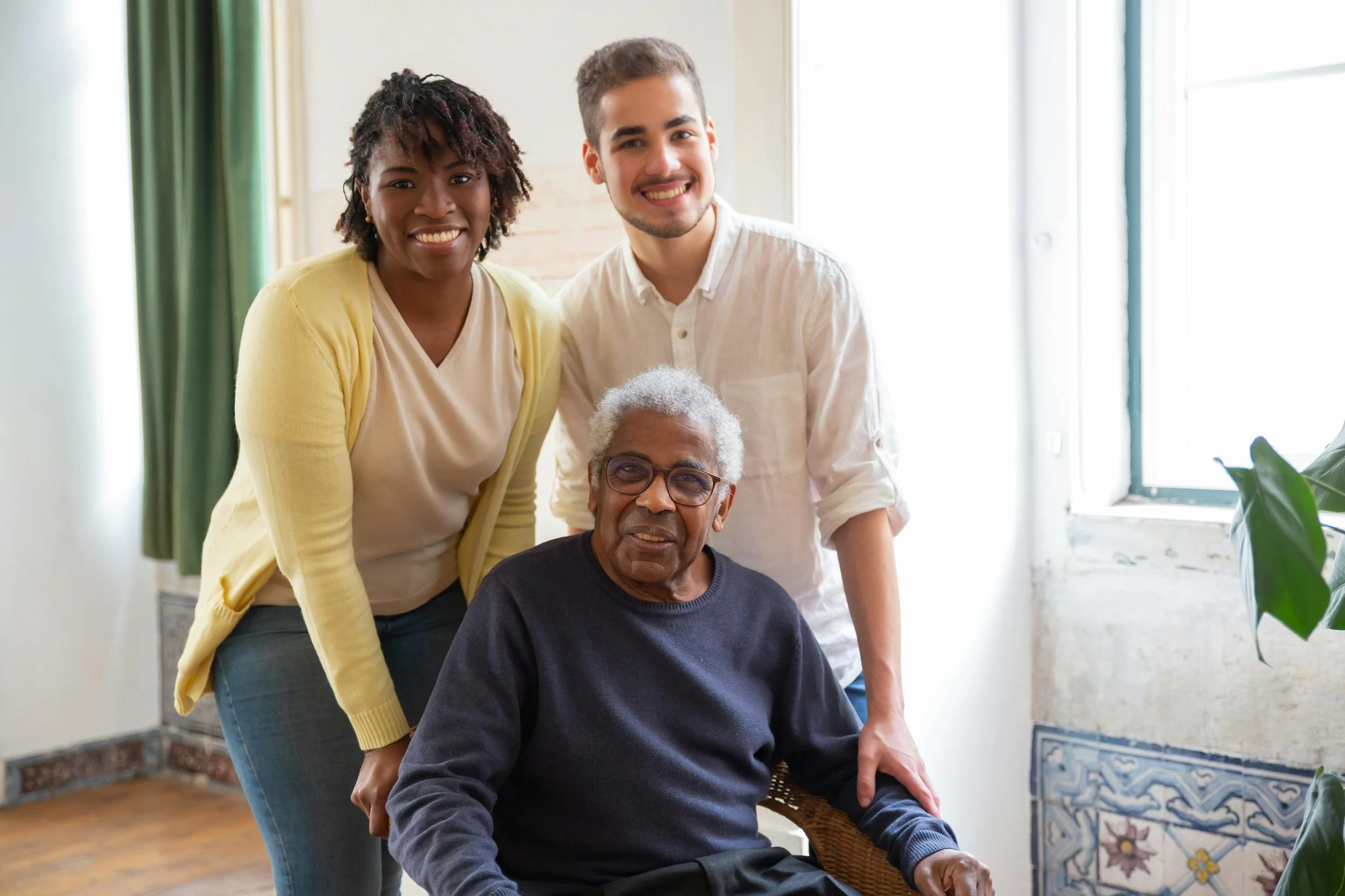 A diverse group of three people, two young adults and an elderly man, gathered together indoors in a well-lit room, smiling for the camera.