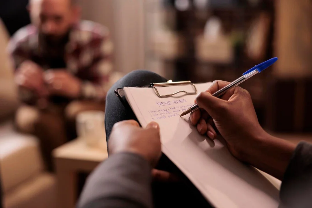 Person taking notes in a notepad during a therapy session, with a blurred man sitting on a sofa in the background.