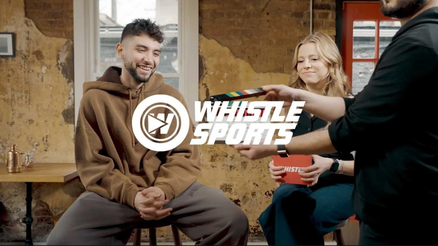 A man in a brown hoodie being interviewed by a woman holding a red box with the WHISTLE SPORTS logo, with a production crew member handing a clapperboard in front of them, in a rustic room with exposed brick walls.
