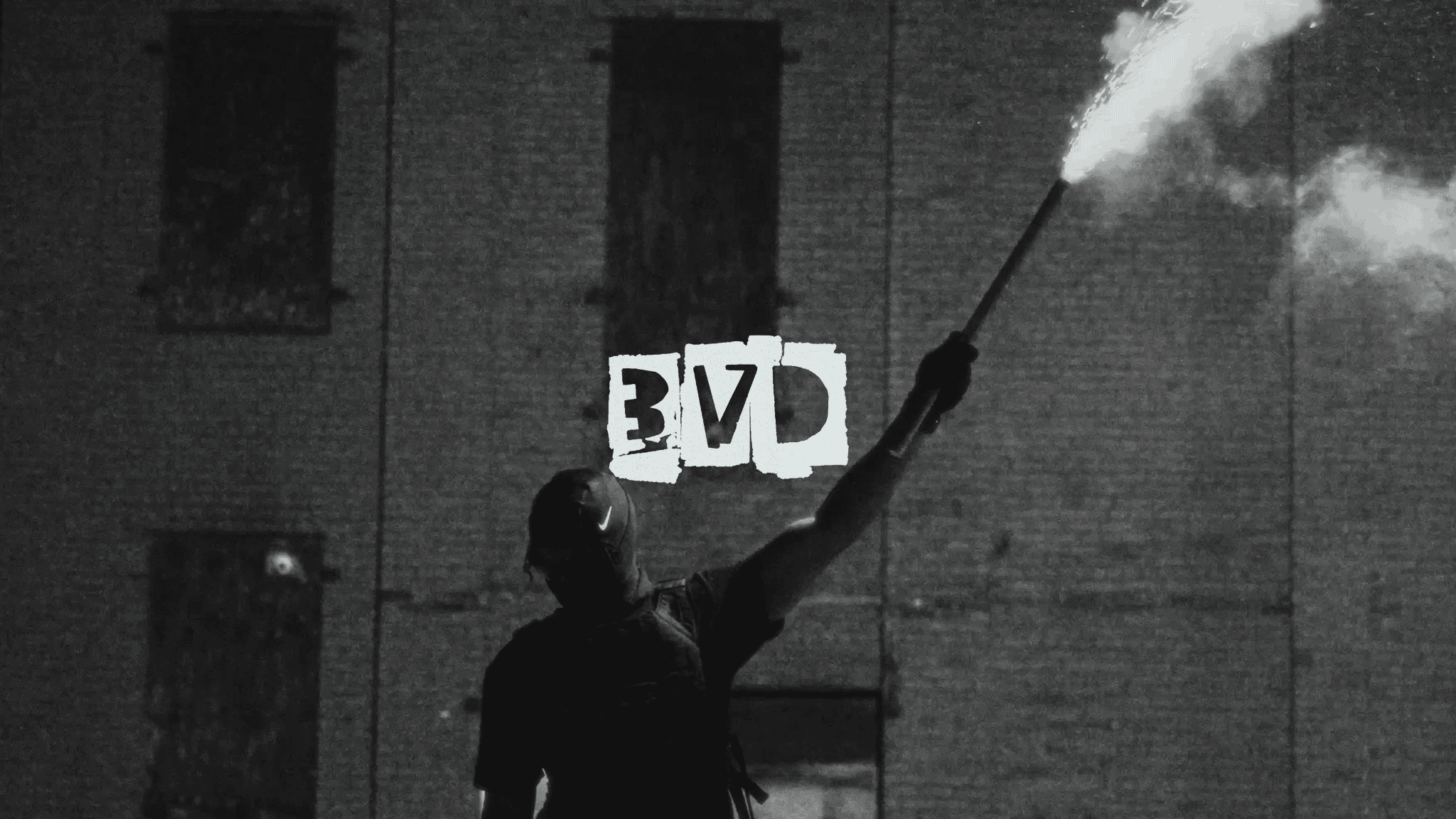 A person wearing a mask and dark clothing holding a lit flare in front of a brick wall. On the wall, there are three blocks with the letters E, V, D.
