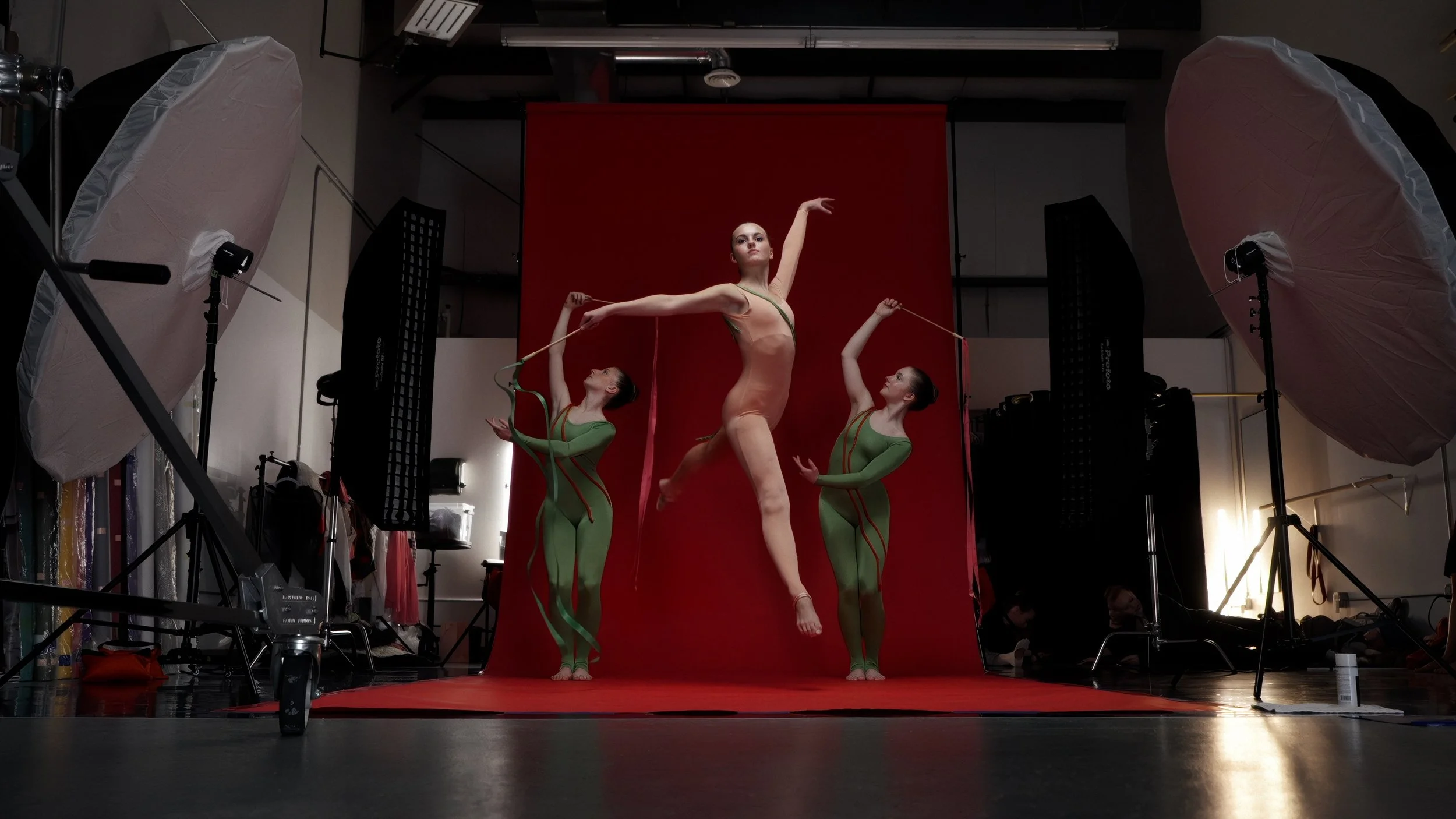 Three dancers perform in a studio with a red backdrop and professional lighting equipment. The central dancer leaps gracefully in a nude-colored bodysuit, while two dancers in green costumes hold ribbons.