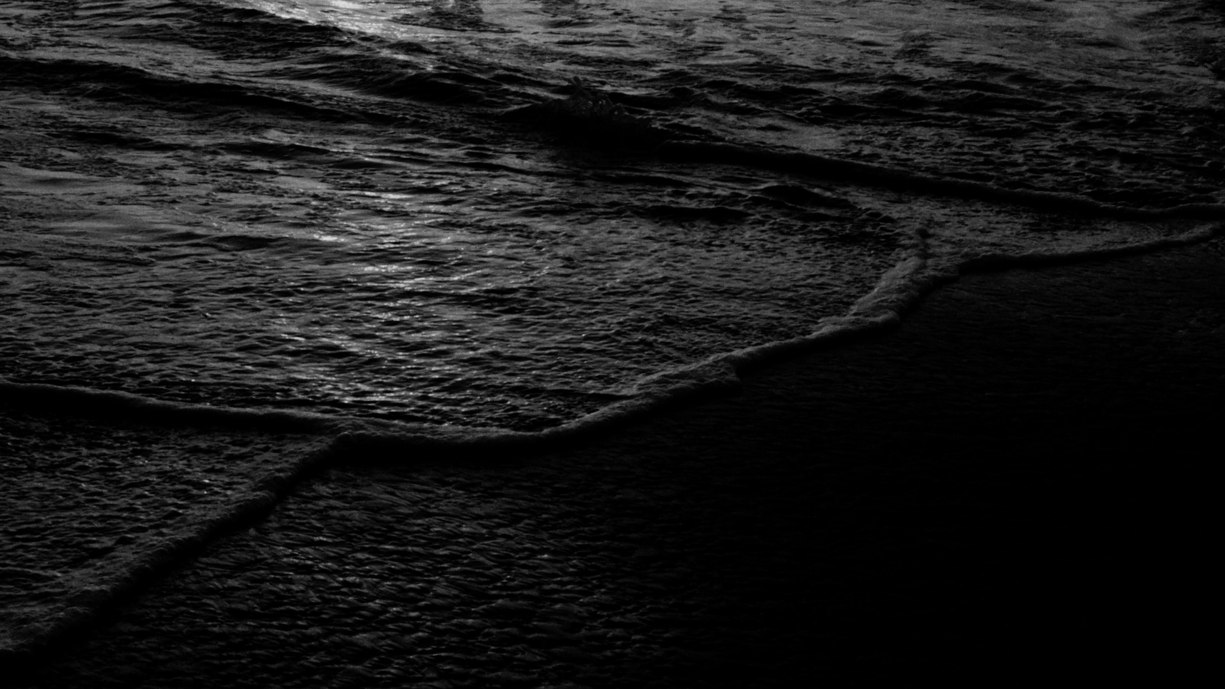 Dark, Pacific Ocean waves gently washing onto a black sandy beach during dusk or dawn.