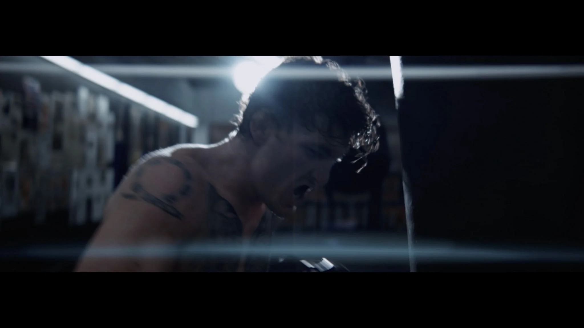 A woman with wet hair and tattoos on her shoulder, likely after a workout, leaning over a punching bag in a gym with a dim, focused lighting environment.