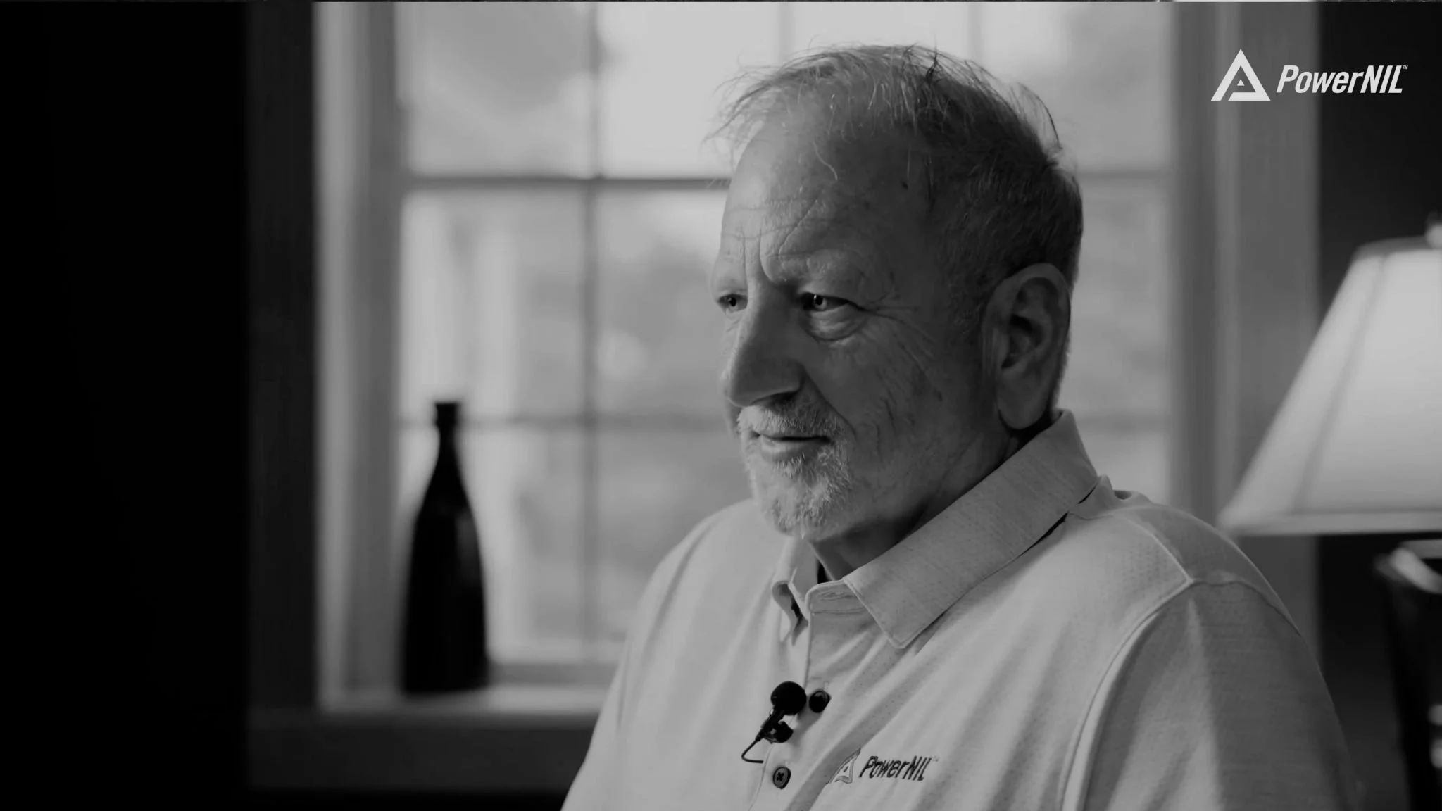 A black-and-white photo of an elderly man with gray hair and a beard, smiling slightly, sitting indoors near a window with a lamp and a bottle in the background, wearing a collared shirt with a small microphone clipped to it.