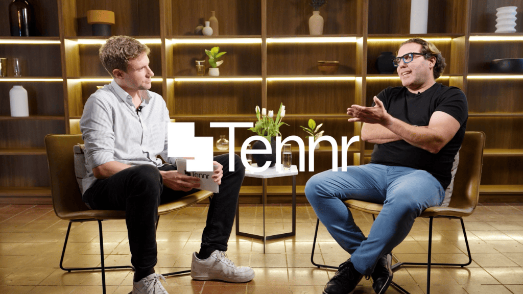 Two men are seated in chairs having a conversation in a modern, warmly lit room with wooden shelving. One man is holding a book, and there are decorative items like plants and vases on the shelves. There is a small table with plants between them, and a logo that says 'Tennr' is overlaid on the image.