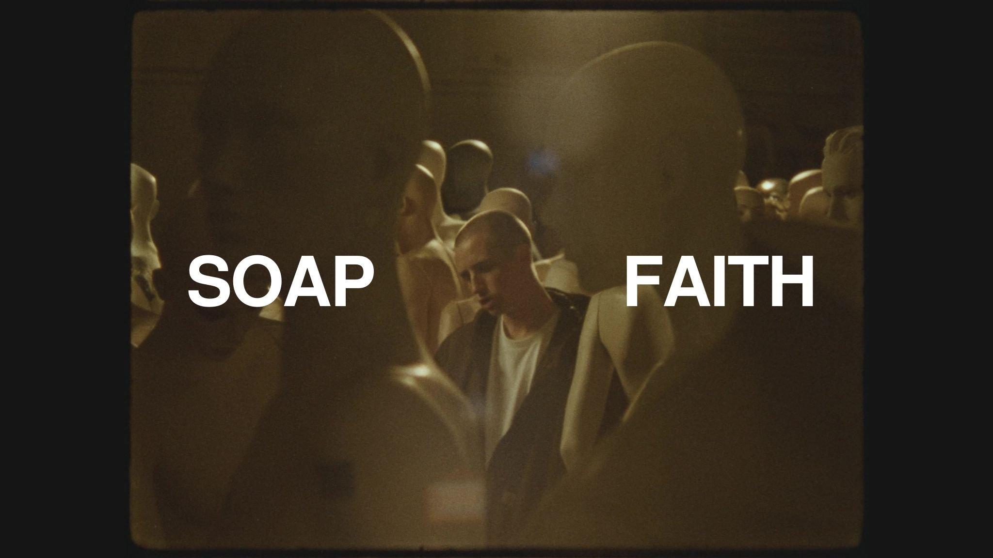 A dimly lit scene with a group of mannequins or wax figures, some wearing masks, standing close together in a crowded space. The words 'SOAP' and 'FAITH' are prominently overlaid on the image, with 'SOAP' on the left and 'FAITH' on the right.