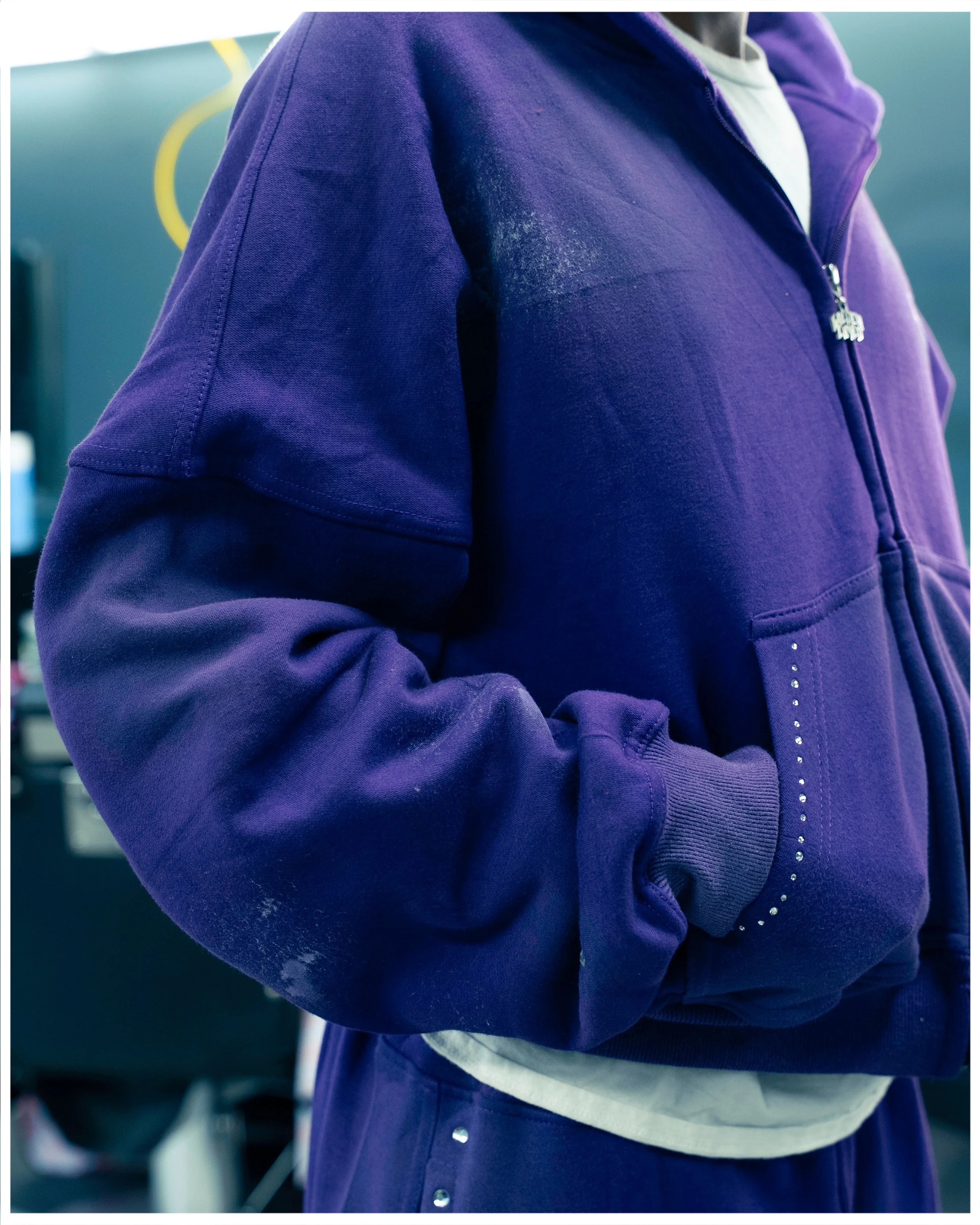 Close-up of a person wearing a purple zip-up hoodie with a white shirt underneath, standing in a setting with blue walls and equipment visible in the background.