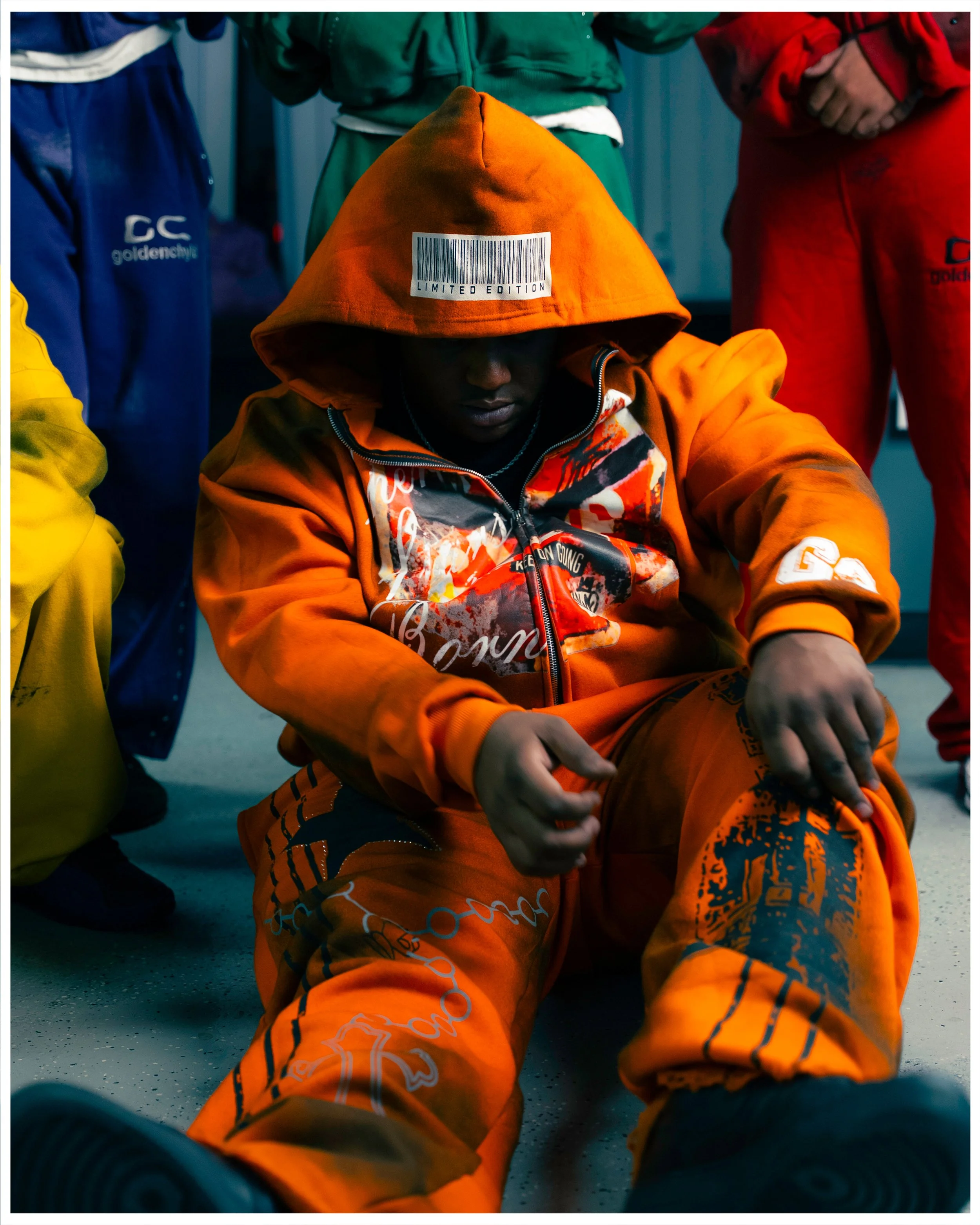 A person dressed in an orange hoodie with the hood over their head, sitting on the floor with their head bowed and hands resting on their knees. Two other people, partially visible, stand around them wearing colorful sportswear.