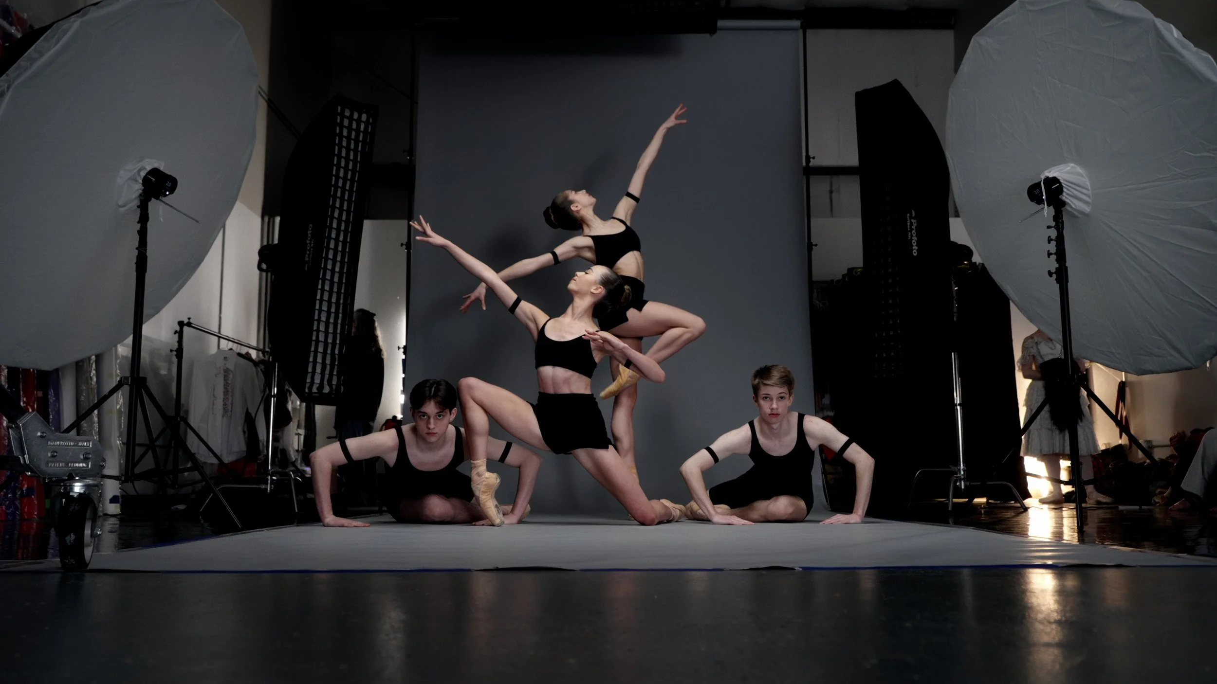 Ballet dancers posed in a studio with large lighting equipment, wearing black outfits and pointe shoes.