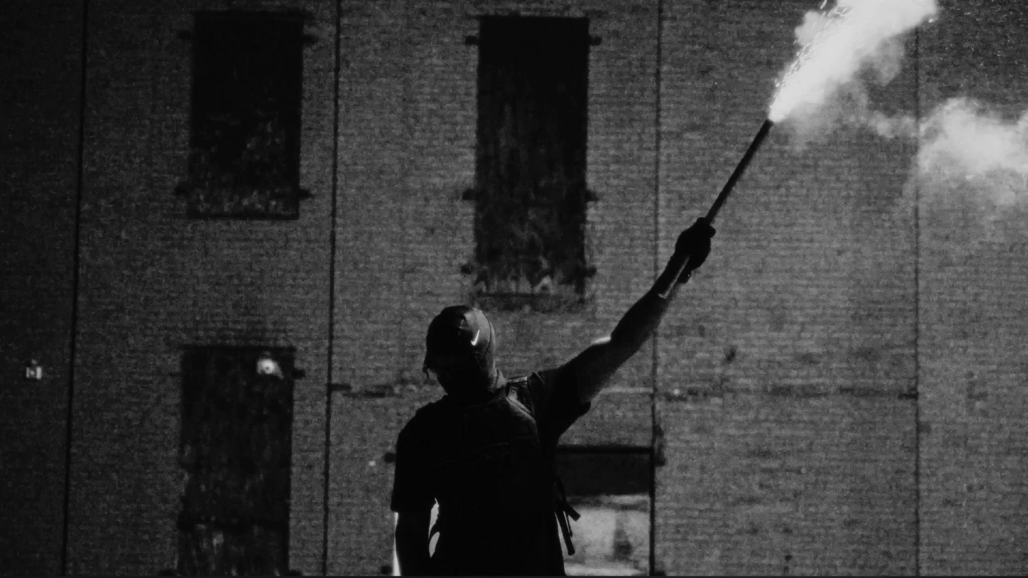 A person with a hood and face covering, holding a flare that emits smoke, in front of a brick building at night.