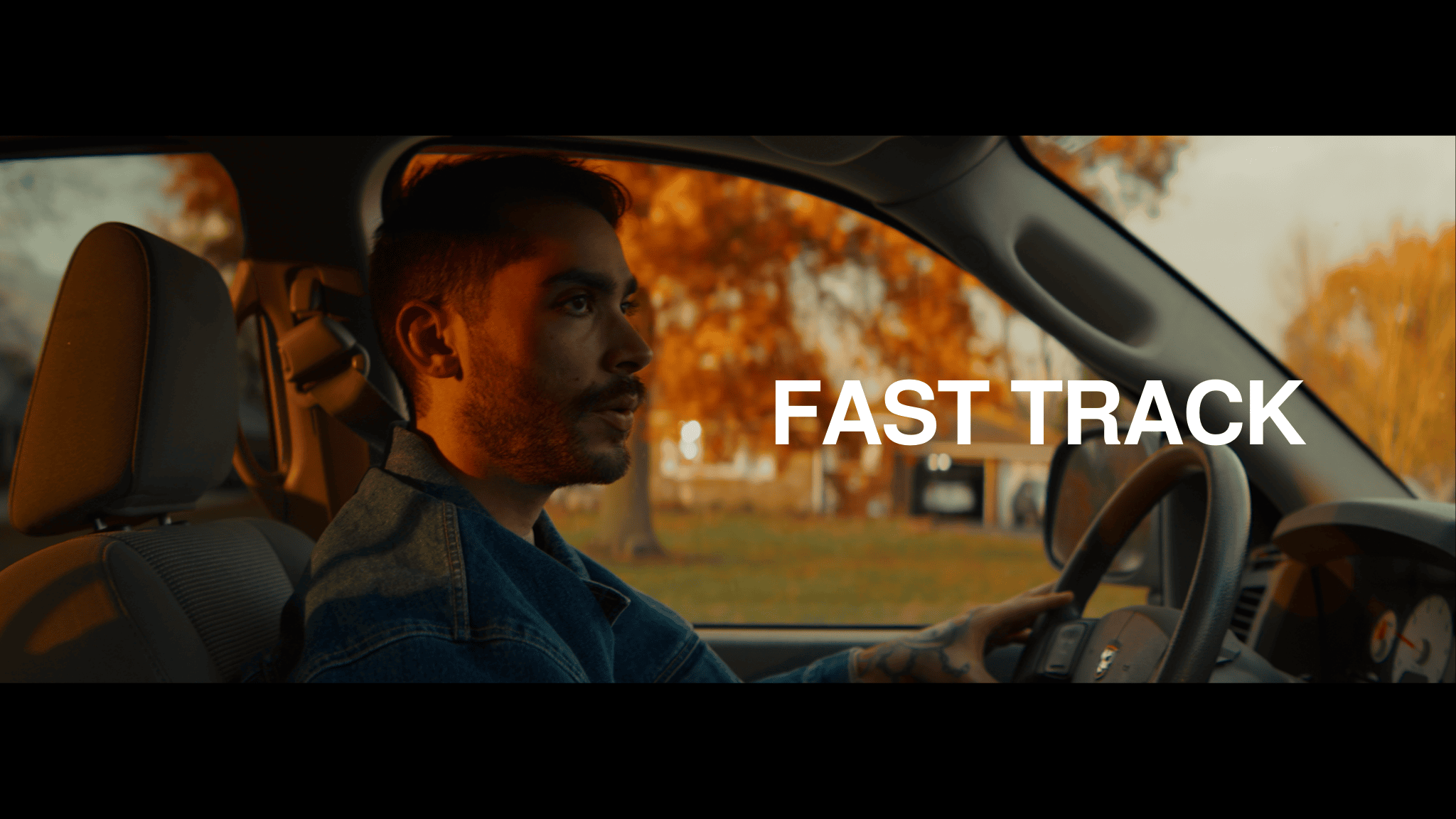 A man with dark hair and tattoos on his hand, wearing a denim jacket, driving a car during autumn with orange and yellow trees outside. The text 'FAST TRACK' is overlaid on the image.