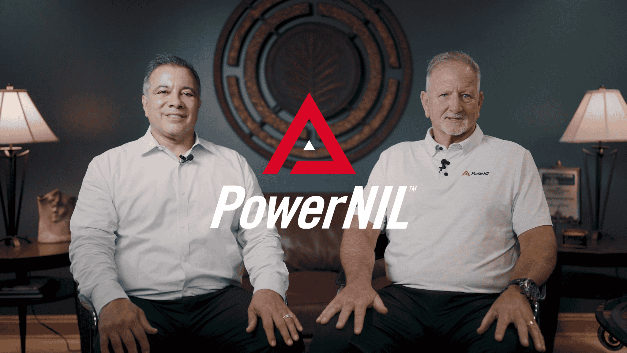 Two men sitting on a couch with PowerNIL logo overlaid in the center. The man on the left is wearing a white shirt, and the man on the right is wearing a white polo shirt with the PowerNIL logo. There are lamps on tables on each side, and a decorative wall art piece behind them.