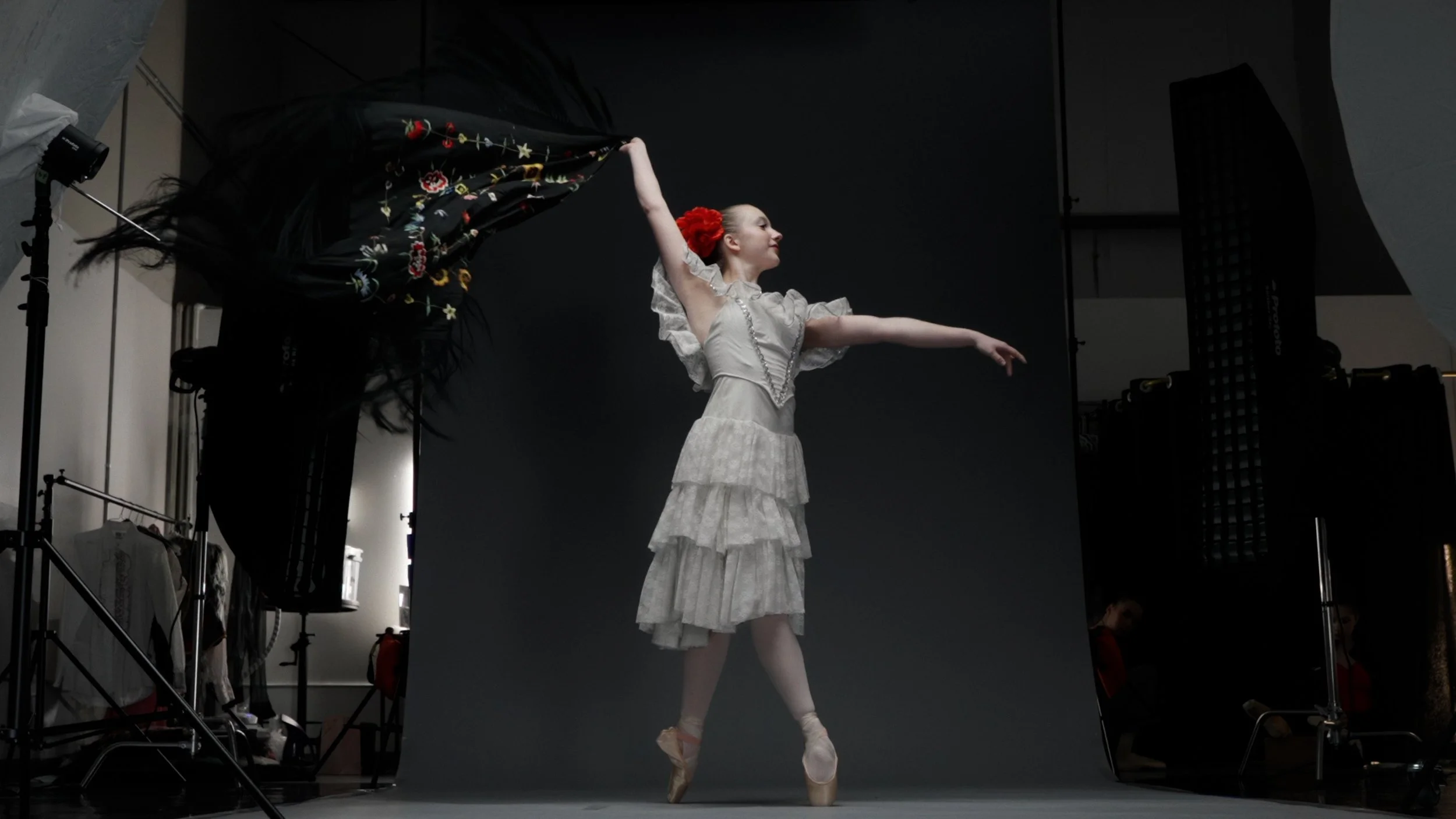 Ballet dancer in a studio, wearing a white tiered dress and red flower in hair, standing on pointe with arms elegantly extended. A floral shawl flutters in motion.